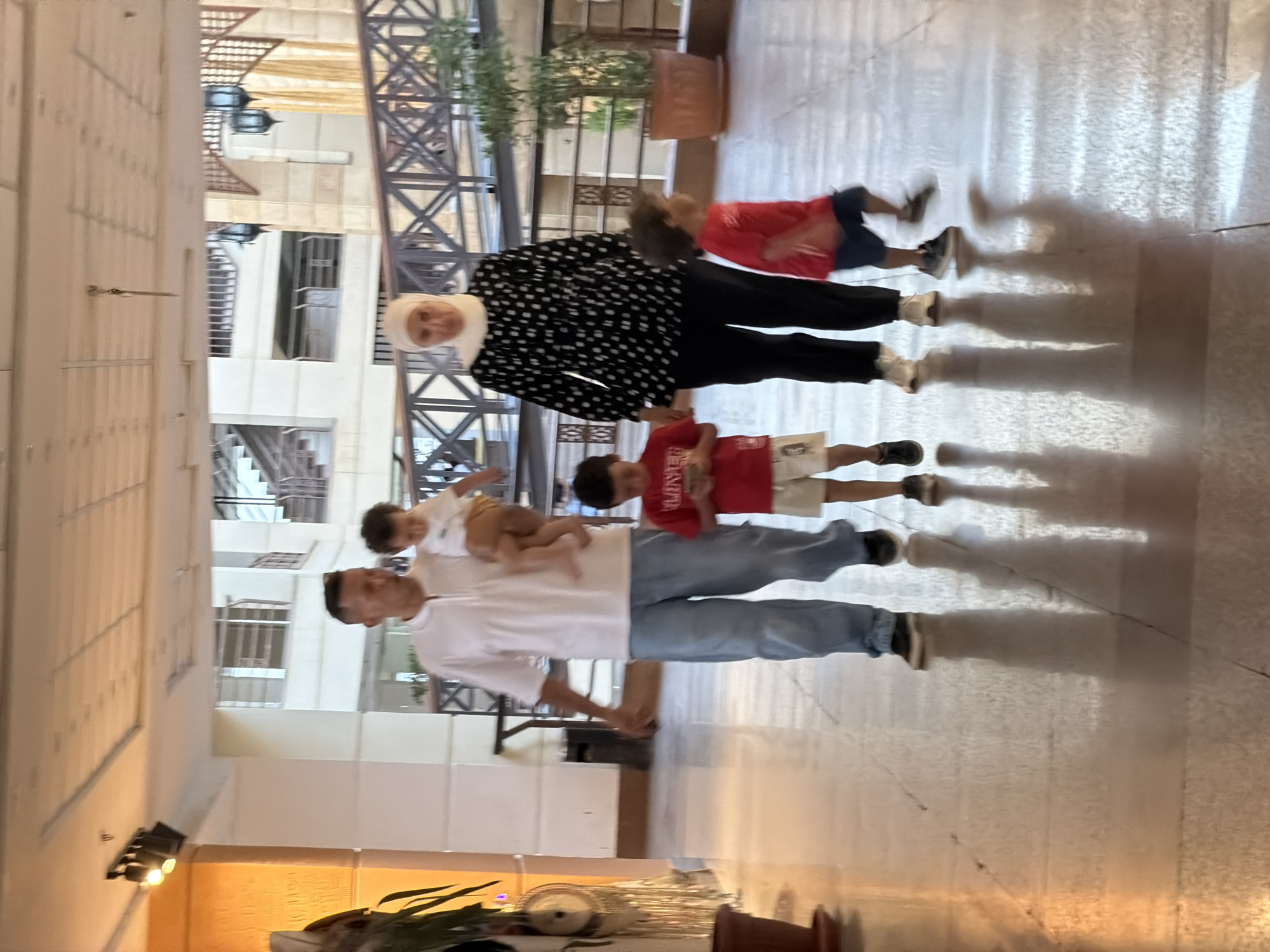 The image depicts a family of five walking in an indoor public space, likely a hotel lobby, in Sharm El Sheikh, Egypt. The father, wearing a white polo shirt and light blue pants, carries a young child in his arms. To his right, a young boy in a red shirt and white shorts looks down at a mobile phone. Behind them, a woman wearing a white hijab and a black and white polka dot blouse walks alongside another young boy in a red t-shirt and blue shorts. The environment features a polished floor reflecting the overhead lighting, large pillars, and architectural elements of what appears to be a resort or shopping complex, including balconies and decorative lamps. The overall lighting suggests an indoor setting with artificial illumination.