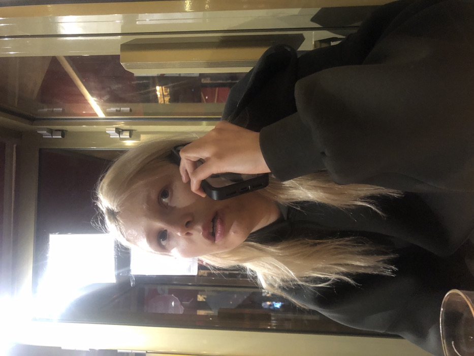 A blonde woman in a black hoodie holds a phone to her ear. She's indoors, possibly in a cafe or restaurant, judging by the window behind her, which reflects the interior. The lighting is soft, with a bright light source behind her. She appears to be in the middle of a phone conversation, with a slightly concerned expression on her face. A glass is visible in the foreground on the right side. The photo is shot at an angle, slightly rotated.