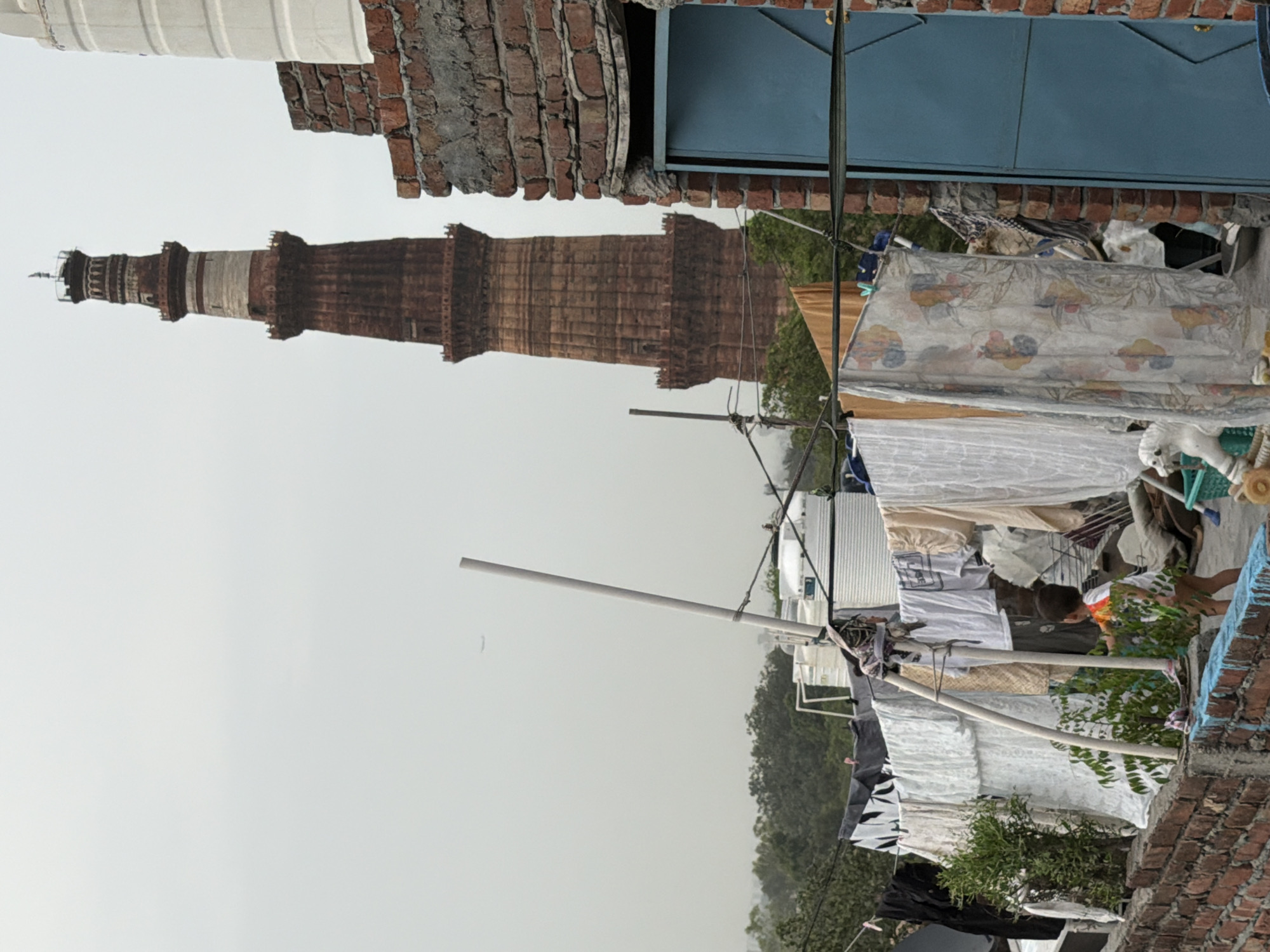 The image depicts an outdoor scene in Delhi, India, with the Qutub Minar, a UNESCO World Heritage Site, visible in the background. In the foreground, a brick wall borders a rooftop area where laundry is hung on clotheslines. A young child, wearing a white and orange shirt and green shorts, is standing near the wall, pointing towards something out of frame. Several water tanks are also present on the rooftop. The sky is overcast, and the overall atmosphere suggests a typical urban environment.