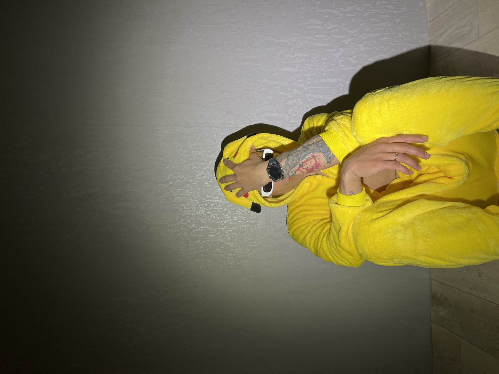 A person is depicted in an indoor setting, viewed from above. They are wearing a bright yellow, fluffy Pikachu kigurumi (onesie) with the character's black eyes and red cheek details visible on the hood, which is pulled over their head. Their head is oriented towards the top of the frame, with the body extending downwards.
The person is lying down, possibly on their back or side, with their right hand (from the viewer's perspective) near their face, holding a pair of white-framed sunglasses. This arm features extensive black and red tattoos. A black smartwatch with a silver bezel is worn on this wrist. Their left hand rests on their chest or stomach area, displaying a simple silver band on one finger.
The background consists of two distinct surfaces meeting at a corner. To the left of the person is a textured, light gray wall, which gradually darkens towards the lower left corner due to shadows. To the right, there's a wooden floor with visible grain. Strong, directional lighting from the upper right casts sharp, dark shadows from the person onto both the wall and the floor.
The scene appears to be set indoors in Chaikovsky, Russia. There is no visible text in the image.