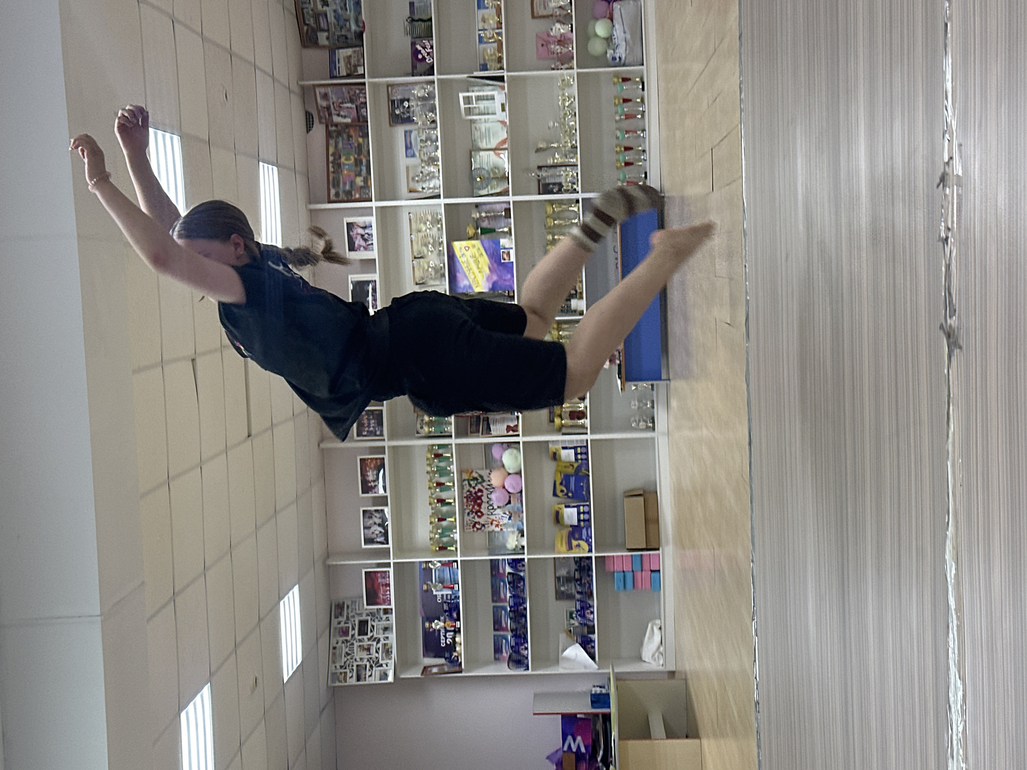 A girl is in mid-air, appearing to jump or dive toward a blue mat. She is wearing a black shirt, black shorts, and striped socks. Her arms are raised above her head. The location appears to be indoors, possibly a gymnastics or dance studio. There is a large shelving unit behind her filled with trophies, pictures, and various objects. The room has white walls with patterned panels. The location is in Votkinsk, Russia.