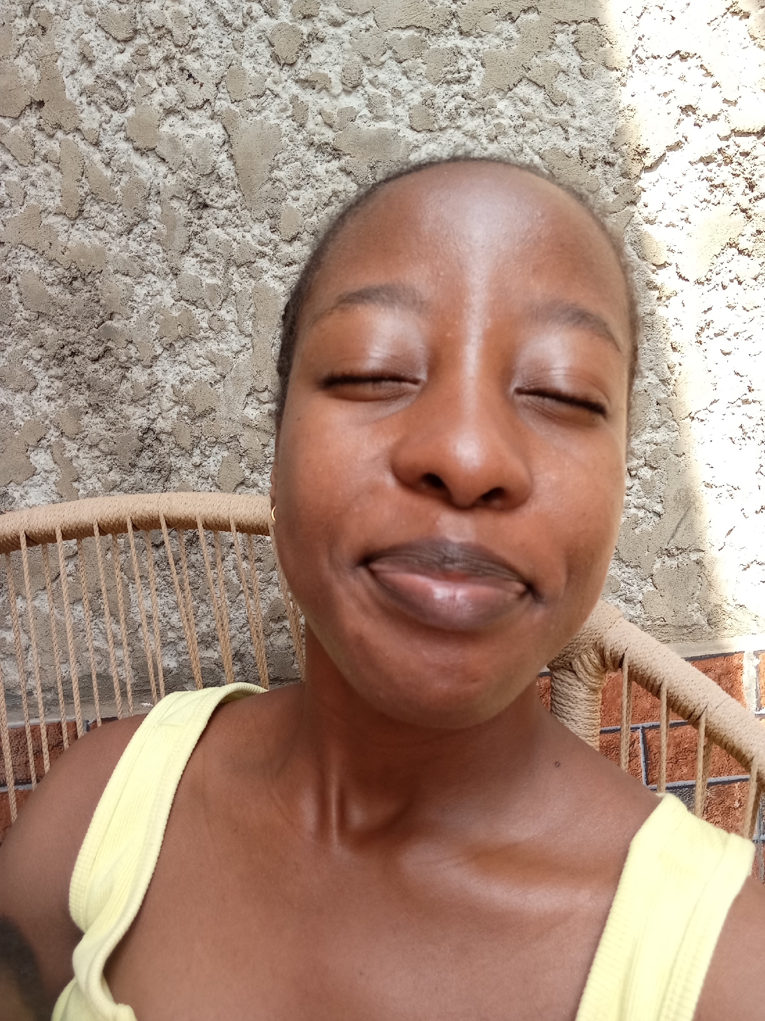 A young Black woman with her eyes closed and a slight smile on her face is pictured in an outdoor setting in Abuja, Nigeria. She is wearing a yellow tank top and a gold hoop earring in her visible ear. She is seated in a rattan chair. Behind her, a textured wall can be seen, with sunlight casting shadows on it. The lighting suggests it is daytime.