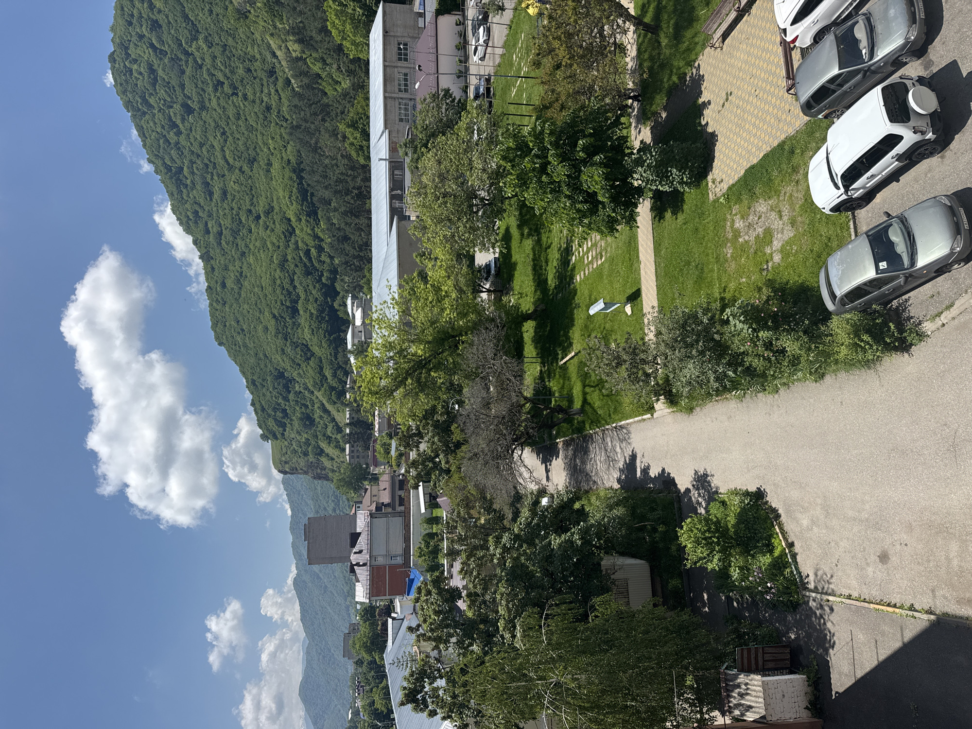 This is a high-angle, full shot of a sunlit scene in Karachayevsk, Russia, featuring a residential area adjacent to a verdant, tree-covered mountain range. In the foreground, to the left, is a small, white brick building with a corrugated metal roof and a wooden door, surrounded by dense green foliage. A paved pathway runs diagonally across the frame from the lower left to the middle right, bordered by grass and trees. Several parked cars, including a white SUV, a silver sedan, and two gray sedans, are visible in the lower right quadrant, on a paved area with yellow patterned tiles.
In the middle ground, a park-like area with green grass and trees is present, with a light blue sign on a pole in the center. Behind the park, several buildings are visible, including a long, light-colored industrial-style building with a metal roof and windows, and other residential buildings with tiled roofs. The background is dominated by a steep, lush green mountainside that rises to meet a bright blue sky with scattered white clouds. The overall impression is a peaceful, somewhat rural setting within a city, with nature prominently featured.