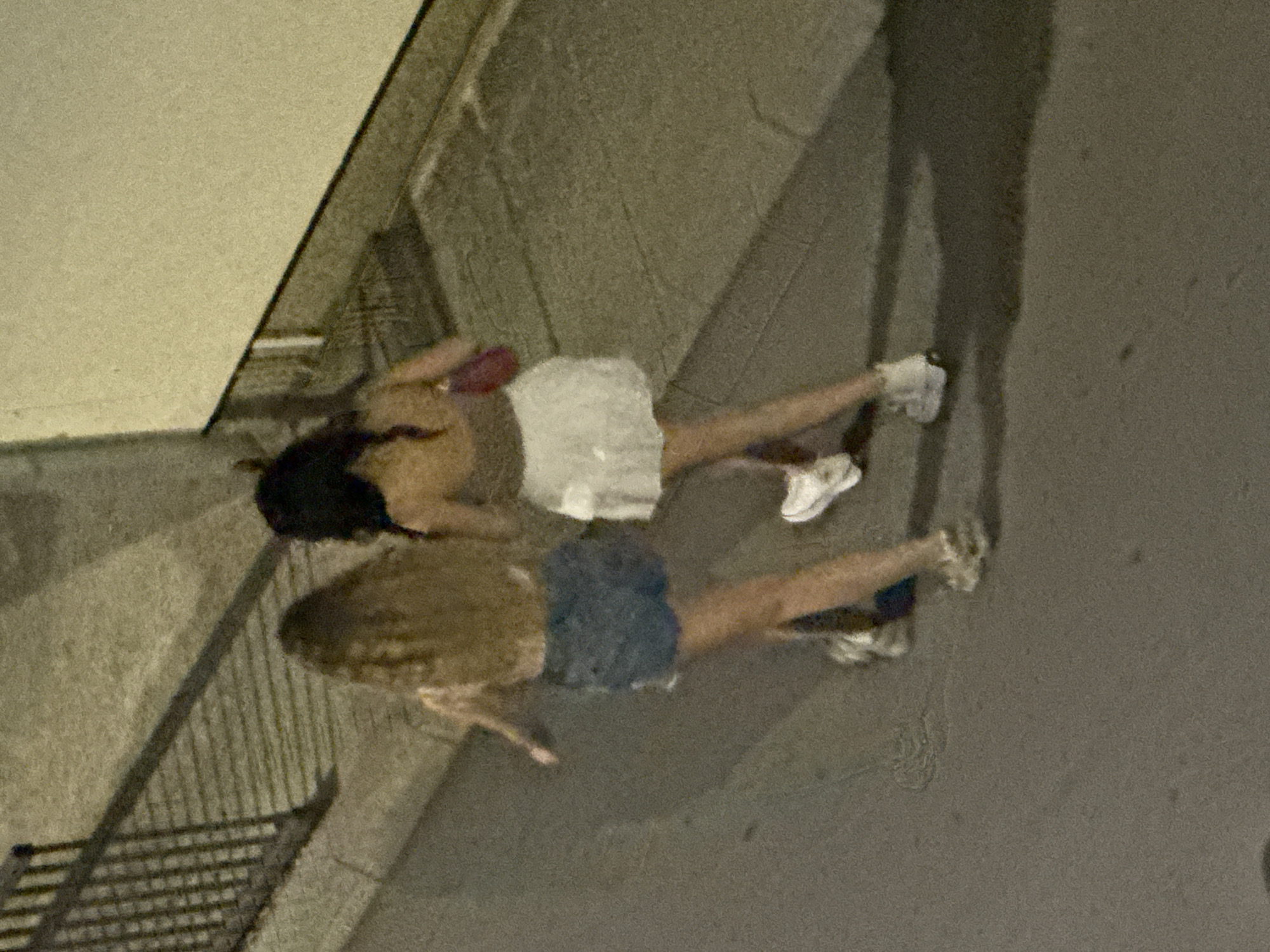 Two young females are walking on a sidewalk next to a street in Budva, Montenegro. The female on the left has long, light brown hair and is wearing denim shorts and white sneakers. The female on the right has dark hair and is wearing a tan top and a white skirt. She is carrying a small red purse. The sidewalk is made of grey concrete with black lines. To the left of the sidewalk is a metal railing. The street appears to be asphalt. The background is a lightly colored wall. The lighting suggests it is nighttime.