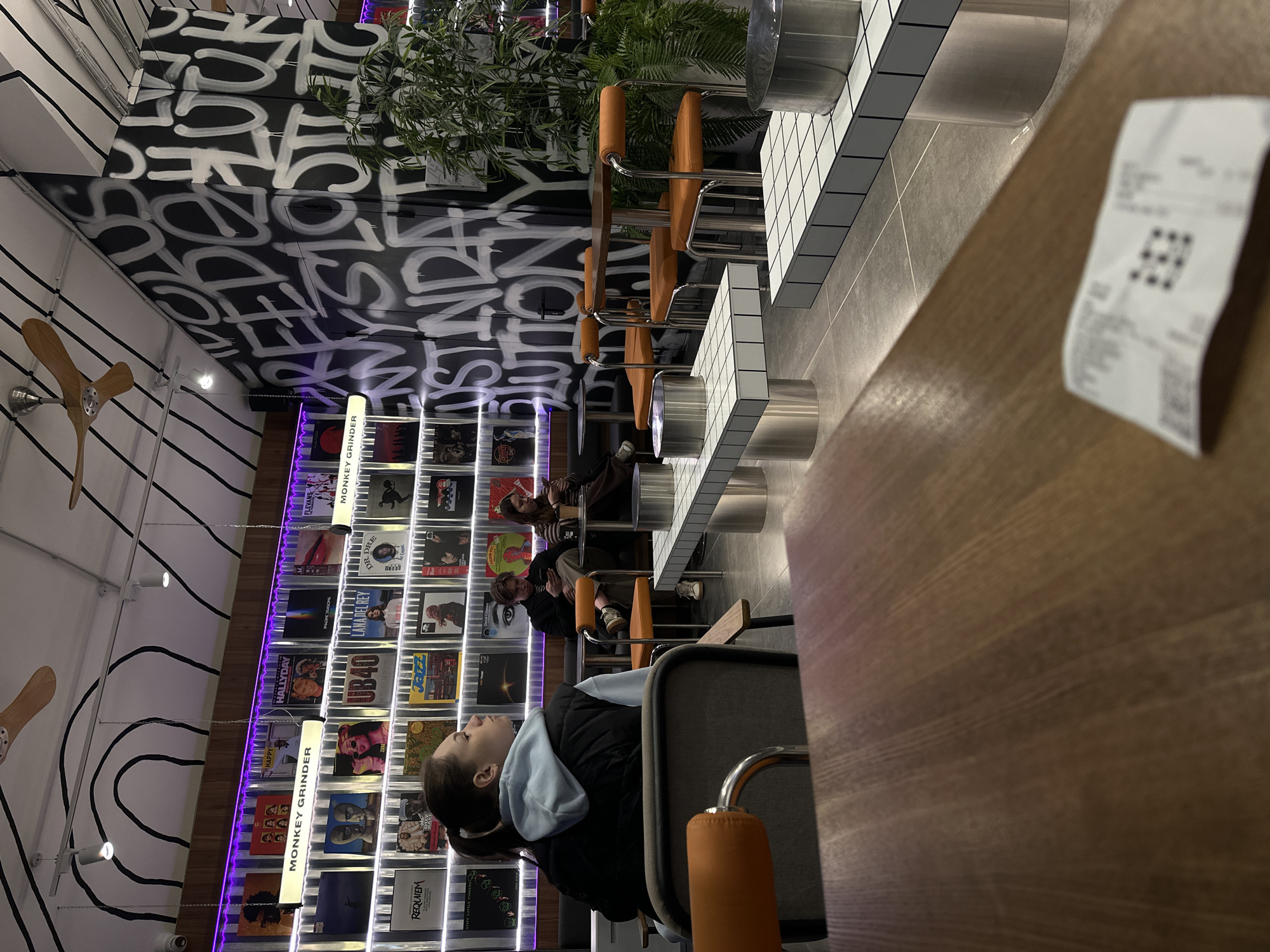 The image shows the interior of "Monkey Grinder," a record store and coffee shop in Perm, Russia. A customer, seen from the side, sits at a counter, potentially waiting for her order. There are other patrons seated in the background, and a receipt sits on a wooden table in the foreground, implying a recent transaction.
The shop's design is notable, featuring an entire wall lined with vinyl record sleeves. Fluorescent lights, some with the store's name "Monkey Grinder" printed on them, illuminate the display. The ceiling has a striking black and white graffiti pattern and an unusual wooden ceiling fan. The counter is adorned with small white tiles.