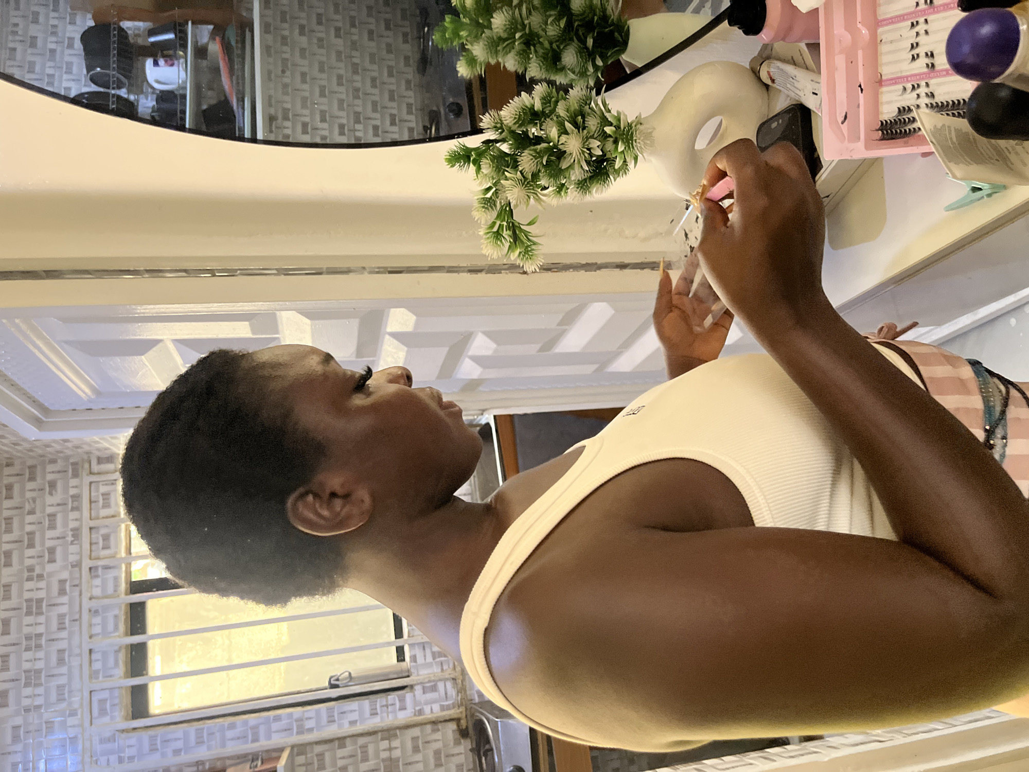 The image shows a young Black woman with short hair, wearing a cream-colored tank top, applying eyelash extensions. She is holding a pair of pink tweezers in her right hand and appears to be focused on her task. Her left hand is also visible, holding the lashes.
In the background, there are several objects on a white surface, including a pink tray filled with artificial eyelashes, a bottle of pink liquid, a white vase with green artificial flowers, and a white mask-like object. There is also a reflection in what appears to be a mirror or a glossy surface, showing more of the room, including what looks like a chair and some shelving with items.
The setting seems to be indoors, possibly a bedroom or a vanity area. The lighting suggests it could be daytime, but it's difficult to determine the exact time of day. The overall mood is one of personal grooming and self-care.
Visible text includes "MIXED CLUSTER EYELASHES" on the pink tray. The scene appears to be set in Abuja, Nigeria, as indicated in the prompt.