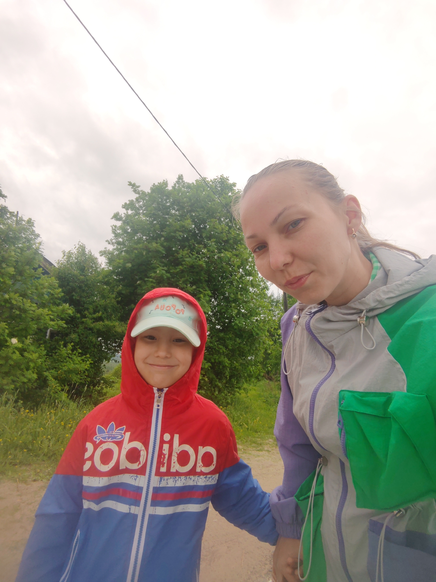 The image is a selfie of a fair-skinned woman and a young child, likely in the vicinity of Чухлэм, Russia, judging by visual cues.
The woman is positioned on the right side of the frame. She has light hair pulled back from her face and is wearing a multi-colored jacket, primarily grey, green, and lavender.
The child, on the left, is wearing a red and blue hooded jacket and a light blue cap.
Both appear to be standing on a dirt or gravel path. The background includes green trees and an overcast sky. There's a power line visible stretching across the upper part of the image. The setting appears to be outdoors, possibly on the edge of a town or rural area. The interaction between the woman and the child suggests a familial connection.