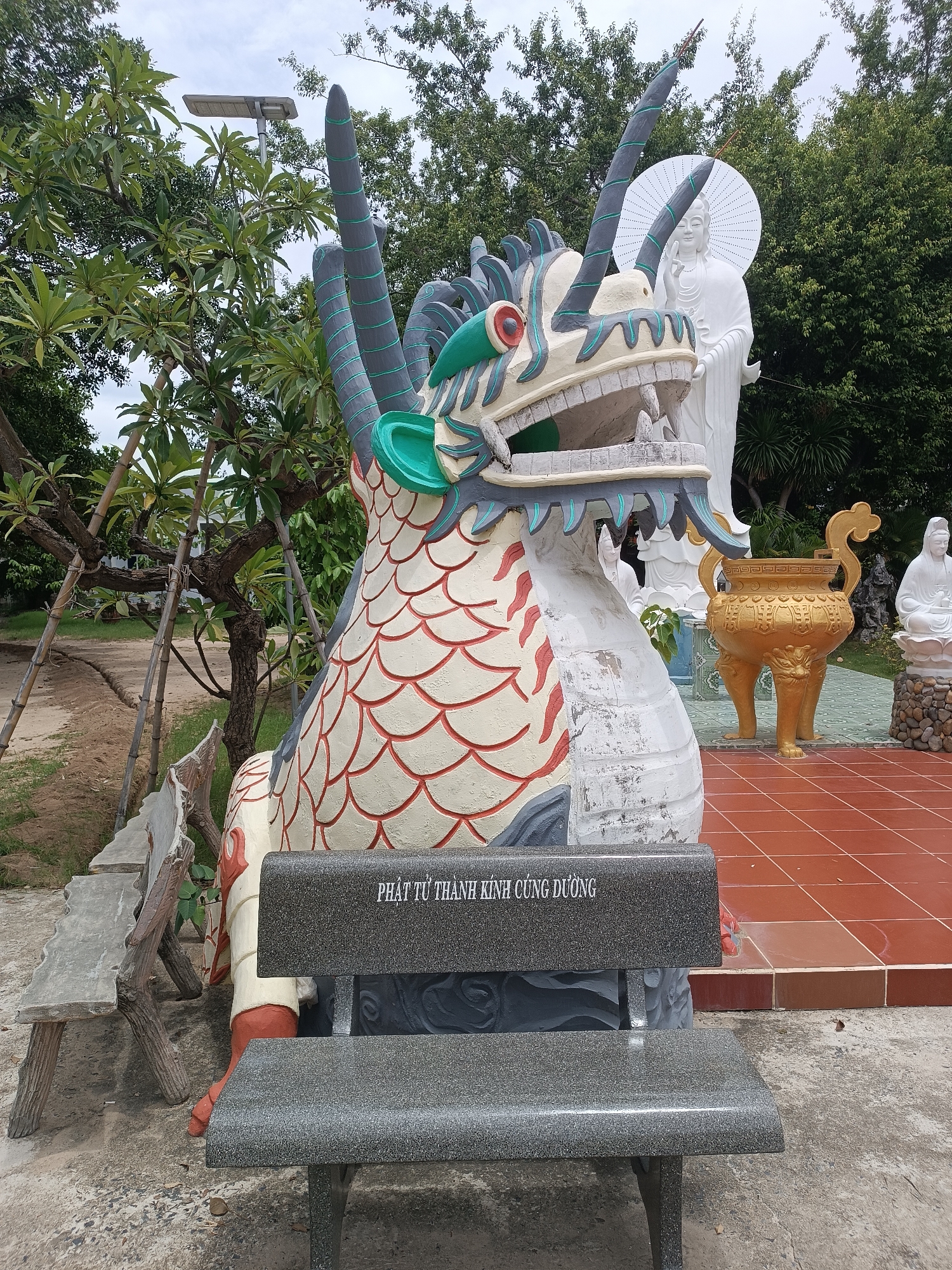 In the city of Cam Ranh, Vietnam, a stone bench sits in front of a large, colorful dragon sculpture. The dragon is predominantly white with red scales outlined in red, and green accents on its head and fins. To the right of the dragon, a white statue of a female figure with a halo stands behind a golden urn. Another smaller white statue is visible further to the right, seated on a lotus flower. In the background, lush green trees and an overcast sky form the environment. Several other stone benches are partially visible to the left of the dragon sculpture. The ground is a mixture of concrete, red tiles, and dirt. The text "PHẬT TỬ THÀNH KÍNH CÚNG DƯỜNG" is inscribed on the backrest of the bench in the foreground.