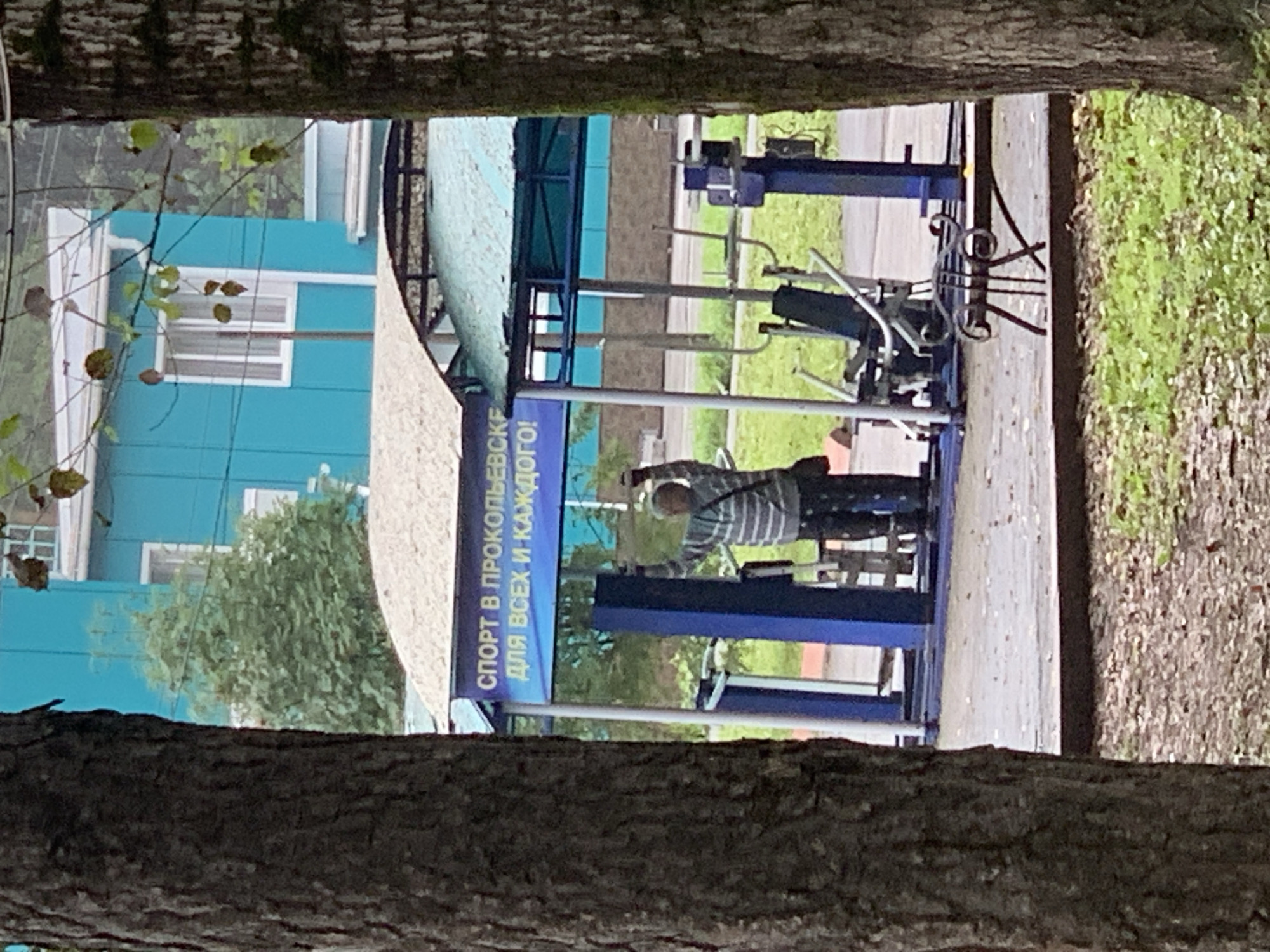 An outdoor gym is located in the city of Prokopyevsk, Russia. A blue and green canopy covers the gym equipment. A sign above the gym reads "SPORT V PROKOPEVSKE DLIA VSEKH I KAZHDOGO!" which translates to "Sport in Prokopyevsk for everyone and each!". A person wearing a striped shirt and pants is exercising on the equipment. The gym is surrounded by trees and a blue building is visible in the background.