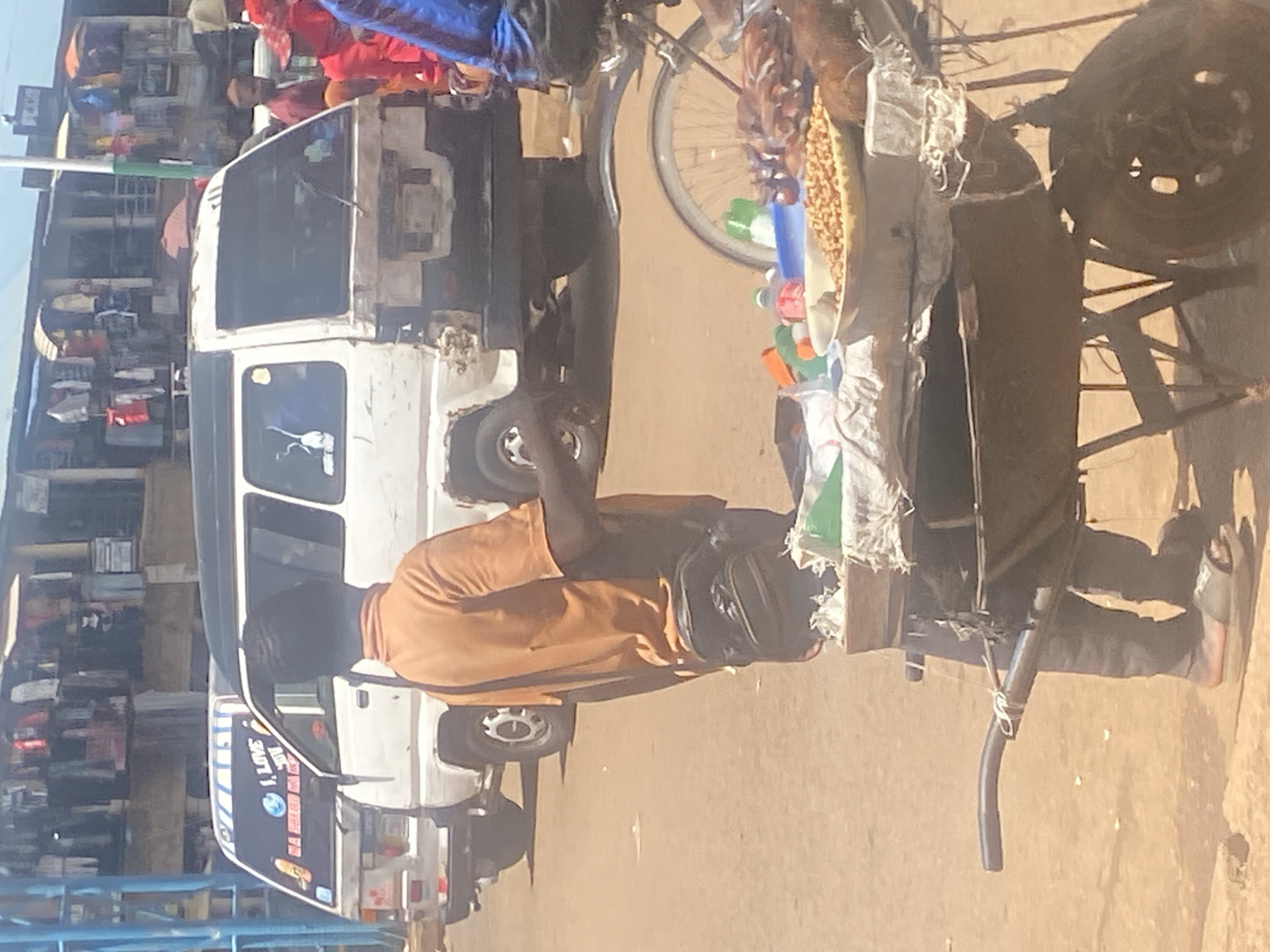 The image depicts a street scene in Bukuru, Nigeria, focusing on a vendor with a cart. A man in an orange t-shirt and dark pants stands behind a cart laden with food items, including a large bowl of nuts and several plastic bottles and bags of goods. He wears a black bag slung across his body and sandals on his feet. The cart itself is a wheelbarrow with a wooden structure on top. In the background, two white vans are parked on a dirt road. The van on the left has "I, LOVE" and a depiction of the earth on its rear window. The background also shows buildings with merchandise displayed outside, suggesting a market environment. The overall lighting indicates it is daytime with bright sunlight.
