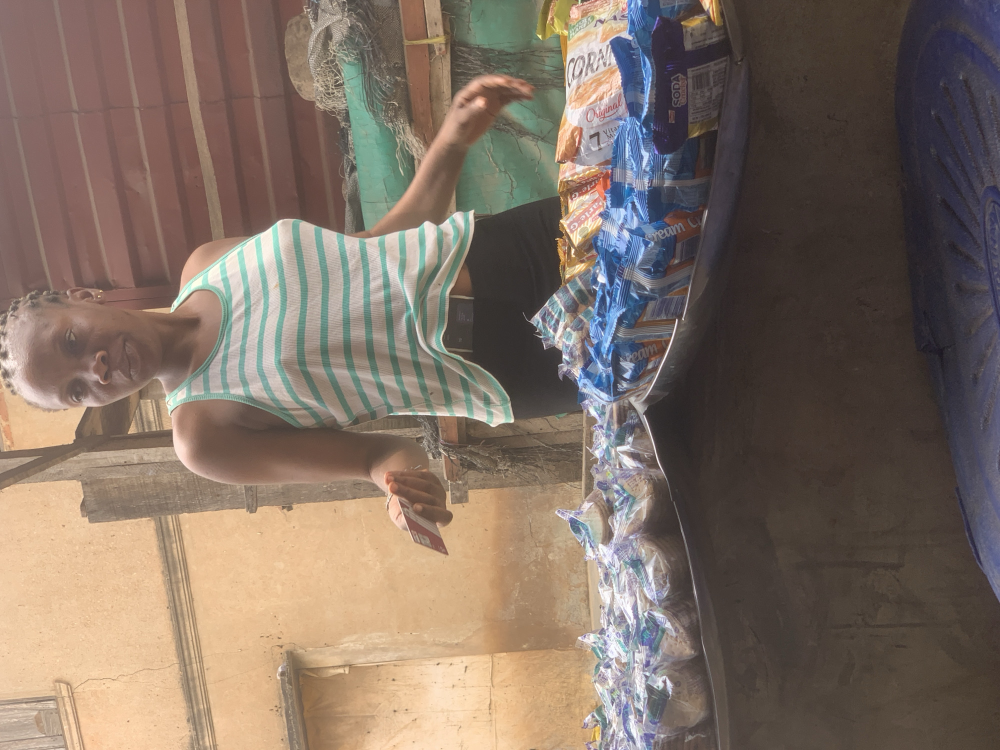 A Black woman is standing behind a stall of food items in Shagamu, Nigeria. She is wearing a white and green striped tank top and black pants. She is holding a red card in her right hand and looking to her left. The stall is covered with various packaged food items, including biscuits and crackers. In the background, there is a building with a textured, light brown wall and a wooden structure. The overall environment suggests an outdoor market setting.