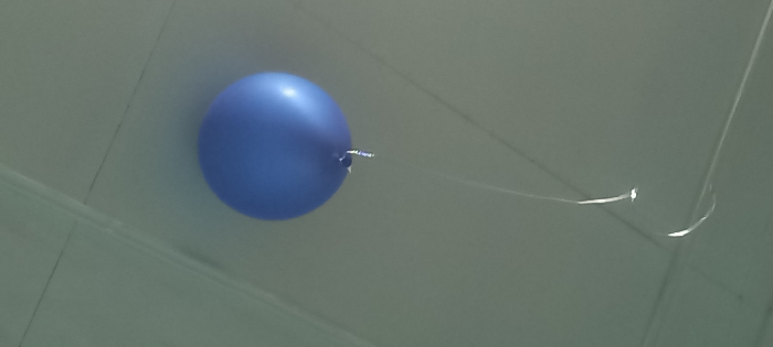A single, inflated, spherical blue balloon is observed floating near a light-colored ceiling. The balloon has a reflective sheen on its upper surface, indicating an unseen light source. A thin, light-colored ribbon or string, appearing white or silver, is tied to the balloon's knot and hangs downwards, curving gently before exiting the bottom-left of the frame. The ceiling surface is light grey or off-white and features linear indentations or seams that suggest a panelized construction, with lines intersecting to form distinct sections. No individuals or additional objects are present in the scene. The image depicts an indoor environment located in Kyzyltepa, Uzbekistan.