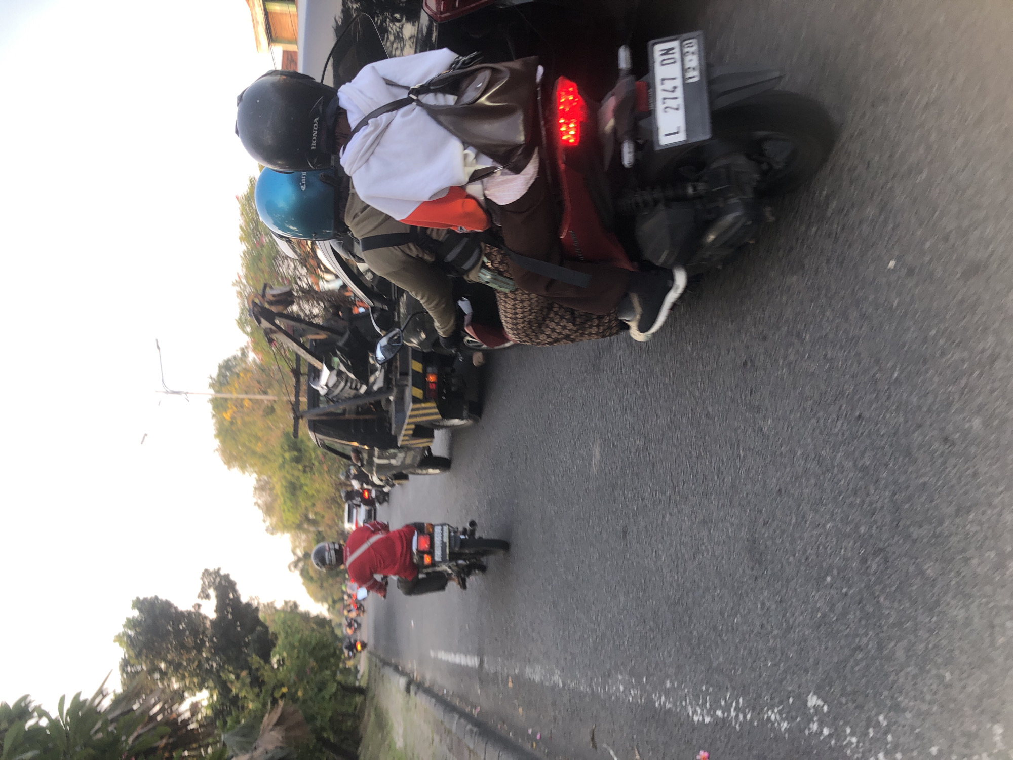 The image depicts a street scene in the city of Gelam, Indonesia, characterized by traffic congestion. In the foreground, a motorcycle carries two passengers, both wearing helmets. The person in the back is wearing a white and orange hooded sweatshirt, and a brown shoulder bag. The driver of the motorcycle is wearing a red shirt and a black helmet. Behind this motorcycle, a tow truck is visible, further indicating a traffic disruption. Several other motorcycles are present in the background, suggesting a typical urban traffic situation. The road is paved, and trees line the sides of the street, with a clear sky overhead. The lighting suggests it is either early morning or late afternoon. The license plate on the motorcycle in the foreground reads "L 2747 DN".