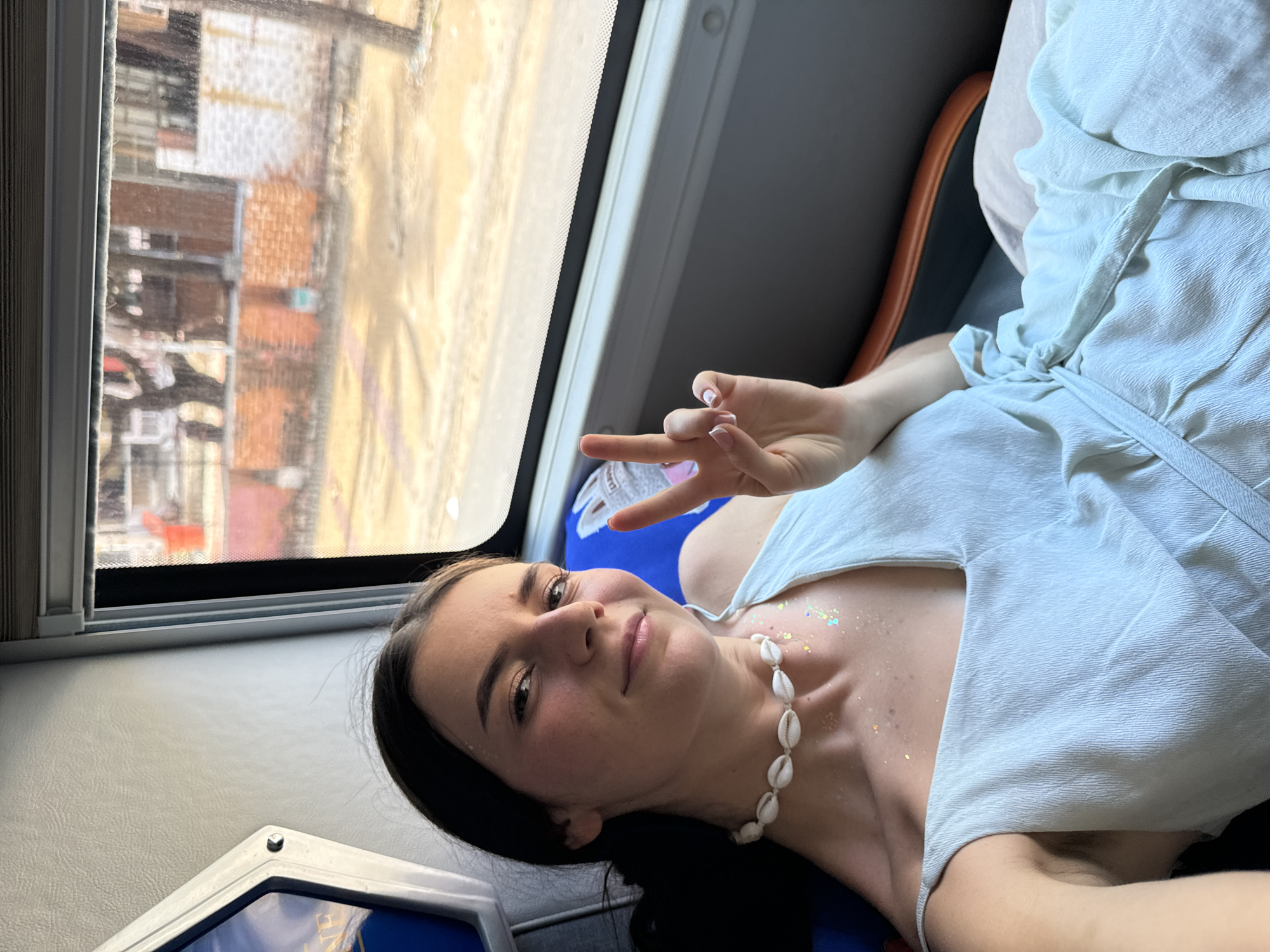 A young woman poses inside what appears to be a moving vehicle with a large window displaying buildings typical of Phu Thuy Ward, Vietnam. The buildings include structures with red-tiled roofs. The woman is reclined, wearing a light-colored dress and a shell necklace, and throwing up a peace sign. There's glitter on her chest. The vehicle has a window and a blue seat.
