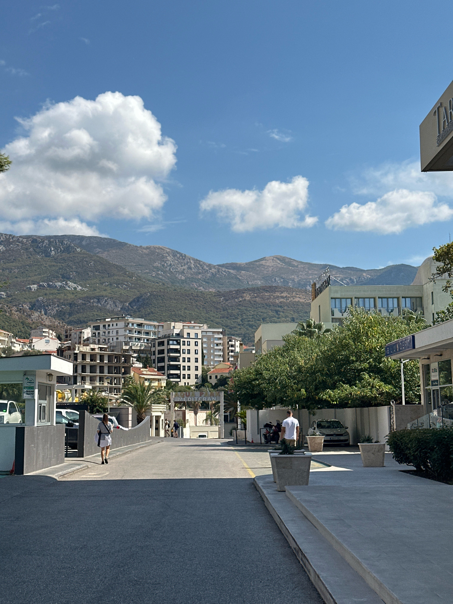 In Bečići, Montenegro, under a bright blue sky with scattered clouds, a street scene unfolds. The foreground features an asphalt road leading towards a security booth on the left, with a person in a grey top and white skirt walking away from it. Adjacent to the road, to the right, is a paved walkway with large square planters. In the middle ground, a row of multi-story buildings with balconies and windows are visible, some appearing under construction. A lush green tree stands prominently in front of one of the buildings. Further back, rolling, tree-covered mountains form the backdrop. A sign with blue lettering, possibly a hotel name, is partially visible on a building in the distance. On the right side of the image, a building with a beige facade has a section of a sign that reads "TAB". In the background, a light-colored car is parked under a covered area. The scene conveys a sense of a developed coastal town with a mix of residential and commercial structures against a natural mountainous landscape.
