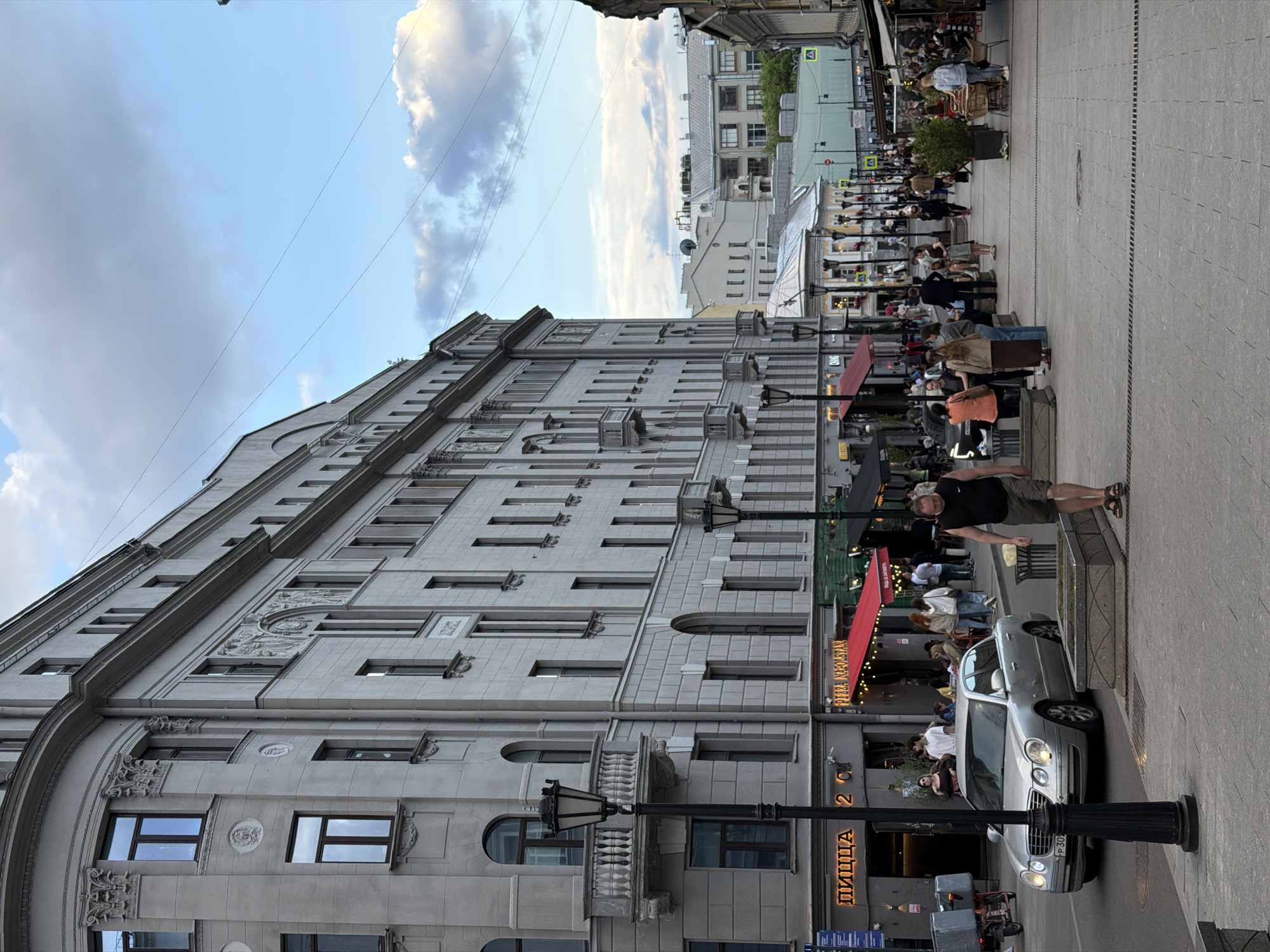 The image captures an urban scene. A tall, grey building with numerous windows occupies a large portion of the left side. The sky above is light blue with scattered white clouds.
On the right, a street bustling with people. There appear to be outdoor seating areas for restaurants or cafes, indicated by red awnings and visible tables and chairs. A man stands near a planter, while a silver car is parked close by. A sign for "ПИЦЦА" (Pizza) is visible on one of the buildings. The scene suggests a lively, pedestrian-friendly area in a city.