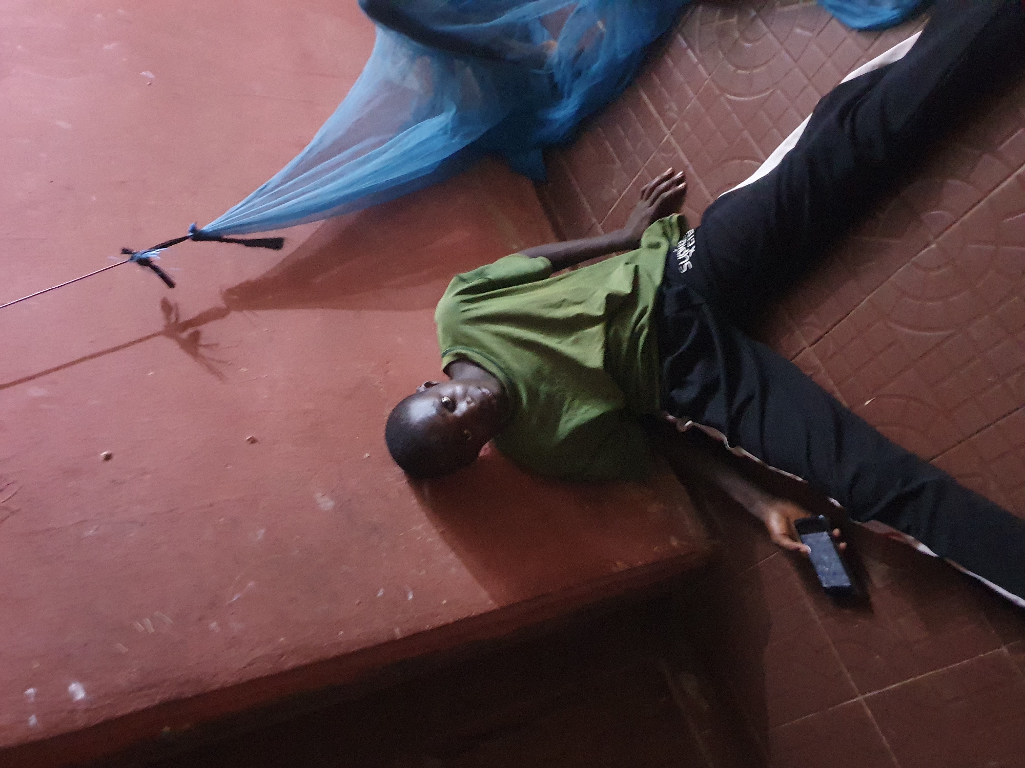 A young person, likely male, with dark skin and short dark hair, lies on their back on a reddish-brown tiled floor, partially resting on a lower reddish-brown platform or bench. They are wearing an olive green short-sleeved t-shirt and black track pants with a white vertical stripe down the side. A dark-colored smartphone is held in their right hand. Above and to the right, a light blue, sheer fabric, resembling a mosquito net, is draped. A thin metal rod or wire is visible, connected to the blue fabric. The overall lighting is dim, indicating an indoor setting, possibly in the evening or a poorly lit room in Idah, Nigeria. The person looks directly at the camera with a neutral expression. Visible text on the left leg of the track pants appears to read "SKATE CRU".