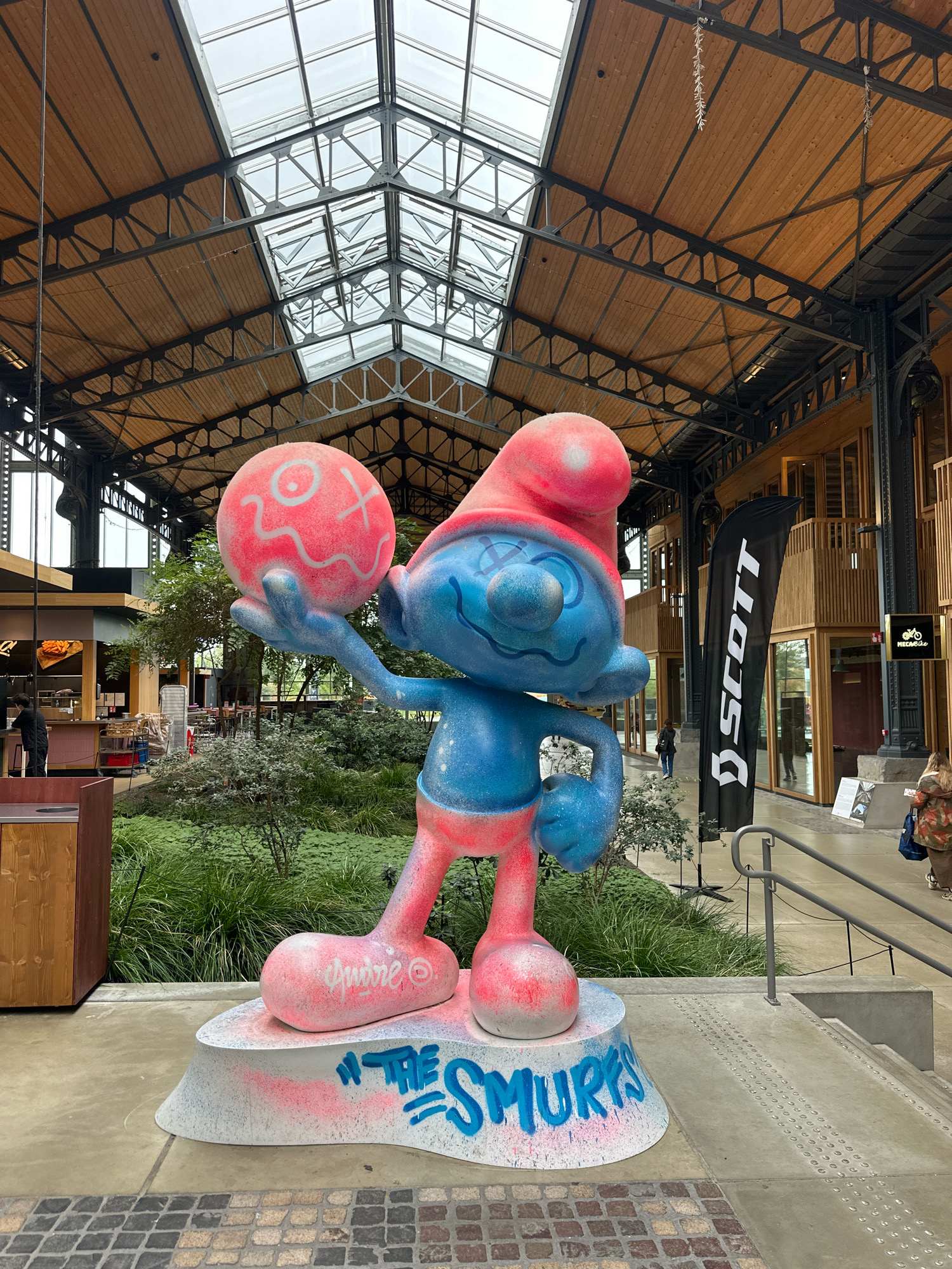 The image shows a large, colorful statue of a Smurf holding a red ball. The statue is blue and red, with pink spray paint on the base and ball. The statue is located in a public indoor space with a wooden ceiling and skylights. There are people and shops in the background. This statue is located in the Docks Bruxsel shopping mall in Brussels, Belgium. A black flag with the "SCOTT" logo is visible behind the statue. The Smurf statue appears to be part of an art installation or exhibition. The floor is paved with tiles and there are stairs with handrails to the right of the statue. Lush green plants are visible behind the statue.