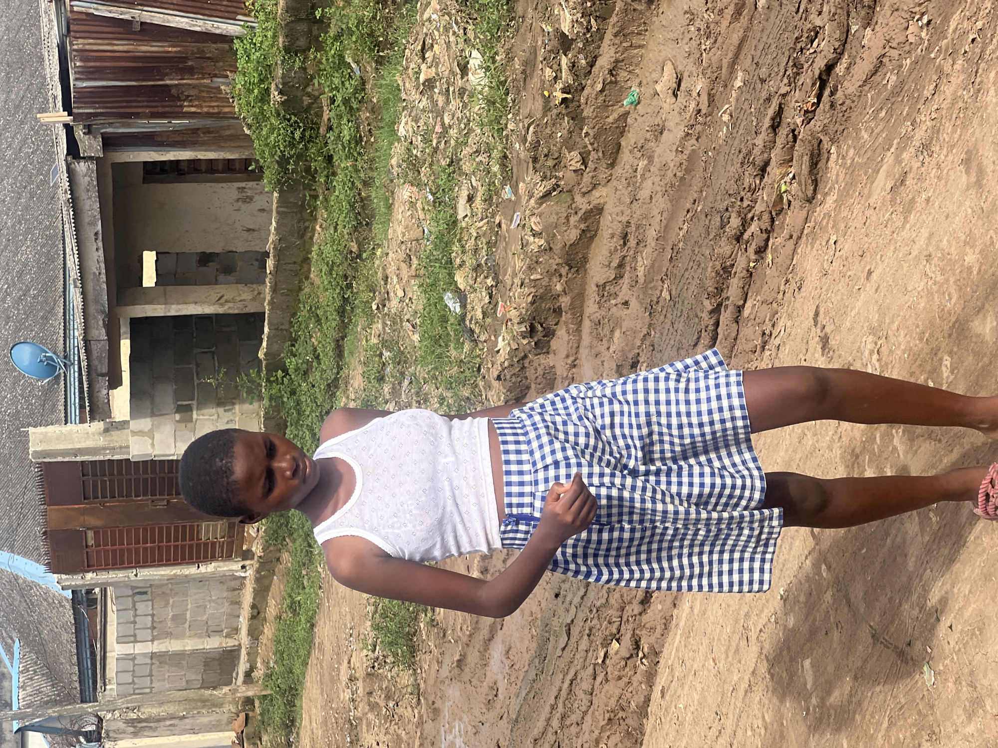A young person stands outdoors in a bright, sunlit environment, likely in Ijegun, Nigeria. The individual has dark skin and short, dark hair, and is wearing a white tank top with a subtle textured pattern and a blue-and-white checkered skirt. Their eyes appear closed or heavily downcast, and one foot with a pink sandal is visible.
The setting is characterized by a dirt ground, which is uneven and appears muddy in places, showing tire tracks and scattered debris or rubbish. Green vegetation, consisting of weeds and grass, grows alongside the muddy path.
In the background, there are several simple buildings constructed from concrete blocks, some appearing unfinished or in disrepair. These structures feature corrugated metal roofs, with one roof having a small blue satellite dish attached. The overall scene suggests a rural or semi-urban area with basic infrastructure, typical of parts of Nigeria. The time of day is clearly daylight, indicated by the strong illumination and visible shadows. There is no visible text in the image.