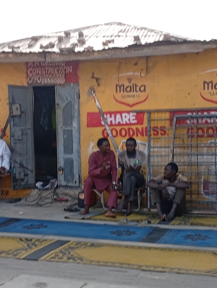 The image captures an outdoor street scene in front of a building with a bright yellow facade and a weathered, corrugated metal roof. The building appears to be a commercial establishment.
Three men are seated casually in front of the yellow wall. On the far left, partially visible, is a person wearing a white garment, seated on a low bench. Next to them, a man in a reddish-brown traditional outfit (possibly a kaftan or agbada) is seated on a small white chair, gesturing with his right hand as if in conversation. He is barefoot. The man in the middle, wearing a light-colored top (possibly grey or beige) and dark trousers, is also seated on a small chair, leaning forward slightly and looking down. A small, rectangular emblem, possibly a flag, is visible on his left sleeve. He is barefoot. The third man, on the right, is sitting directly on the ground, cross-legged, with his arms resting on his knees, looking towards the left. He is dressed in a light top and dark trousers and appears to be barefoot or wearing minimalist footwear.
The yellow wall of the building features several visible signs. Above an open, dark metal double-doorway, a red sign with white text reads "ATA BEDING CONSTRUCTION" (likely "ATA BUILDING CONSTRUCTION") and below it, a phone number "0908xxxxxx". To the right of the door, the "Malta GUINNESS" logo is prominently displayed twice. Below one of these logos, a red banner with white text reads "SHARE GOODNESS." A long, light-colored pole leans against the yellow wall, extending upwards. To the right of the seated men, a large metal grid or rack stands upright, partially obscuring the "SHARE GOODNESS" banner. A small number "107" is visible on the wall to the left of the doorway, near the bottom.
On the ground in front of the men, a blue mat with a decorative pattern is laid over a larger yellowish-brown patterned surface, possibly another mat or paved area. Electrical wires and cables are scattered on the ground near the open doorway, suggesting some form of work or activity involving electronics or power tools might be ongoing inside or around the shop.
The scene is lit by natural daylight, indicating it is daytime. The sky is bright and appears overcast or hazy. The overall atmosphere is relaxed, with the men appearing to be conversing or simply resting in front of the shop. The visible branding and the style of attire suggest the location is in an African country, likely Nigeria.