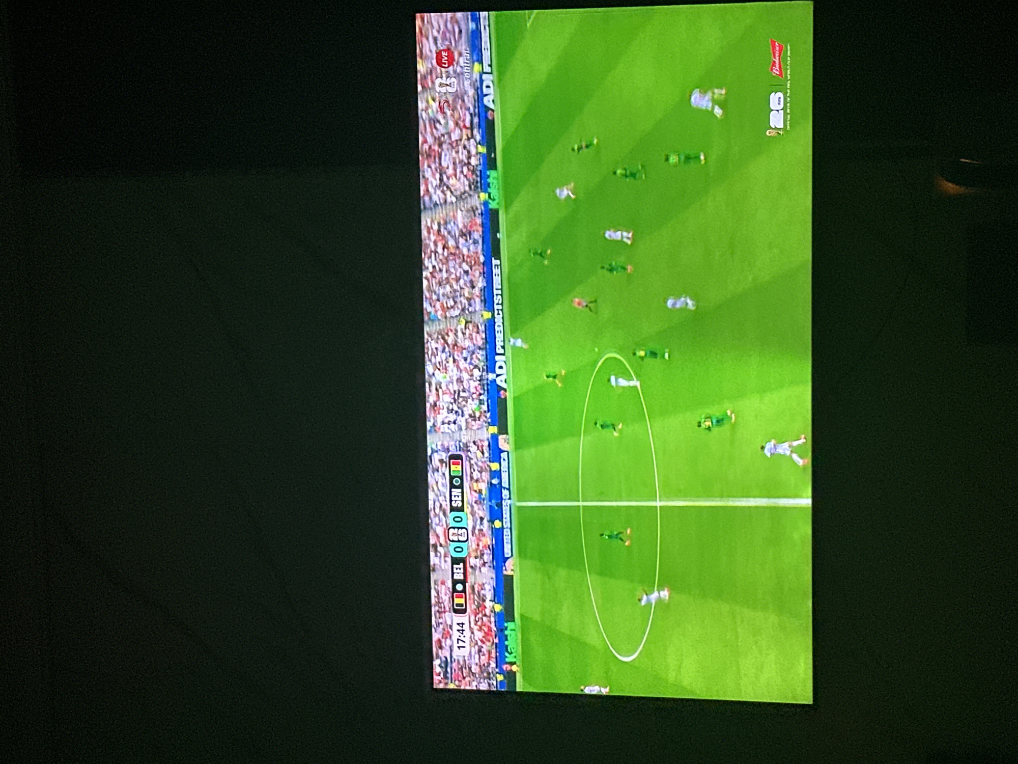 The image displays a television screen showing a soccer match between Belgium (BEL) and Senegal (SEN). The score is 0-0, and the game is at the 17:44 mark. The screen is framed by a dark border, suggesting it is a television within a room. The soccer field is bright green with white lines marking the boundaries and center circle. Several players in white and green uniforms are visible on the field. In the background, a stadium filled with spectators wearing various colored clothing can be seen. Advertising banners are present along the sideline. The scene is captured in Gwarinpa, Nigeria, indicating the television is located in that city.