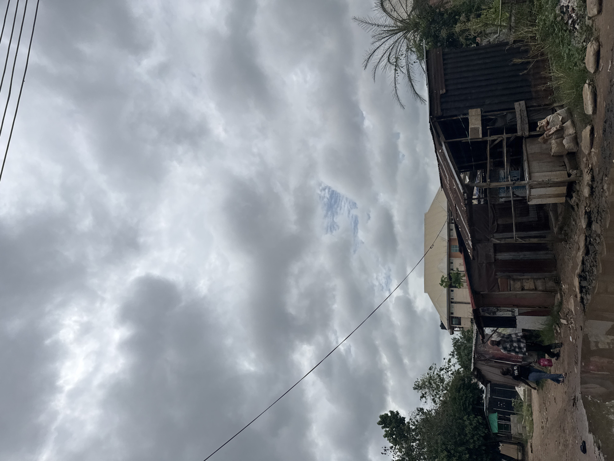 The image depicts a street scene in Ihiagwa, Nigeria, under a heavily overcast sky. A dilapidated corrugated metal structure stands to the right of the frame, with evidence of makeshift construction and various objects stored within. To the left, a taller building can be seen partially obscured by the metal structure. At least three people can be seen walking along the unpaved road. The road appears to be wet, possibly due to recent rain. There is vegetation and a palm tree visible. Electrical wires stretch across the sky. The environment suggests a low-income or underdeveloped area.