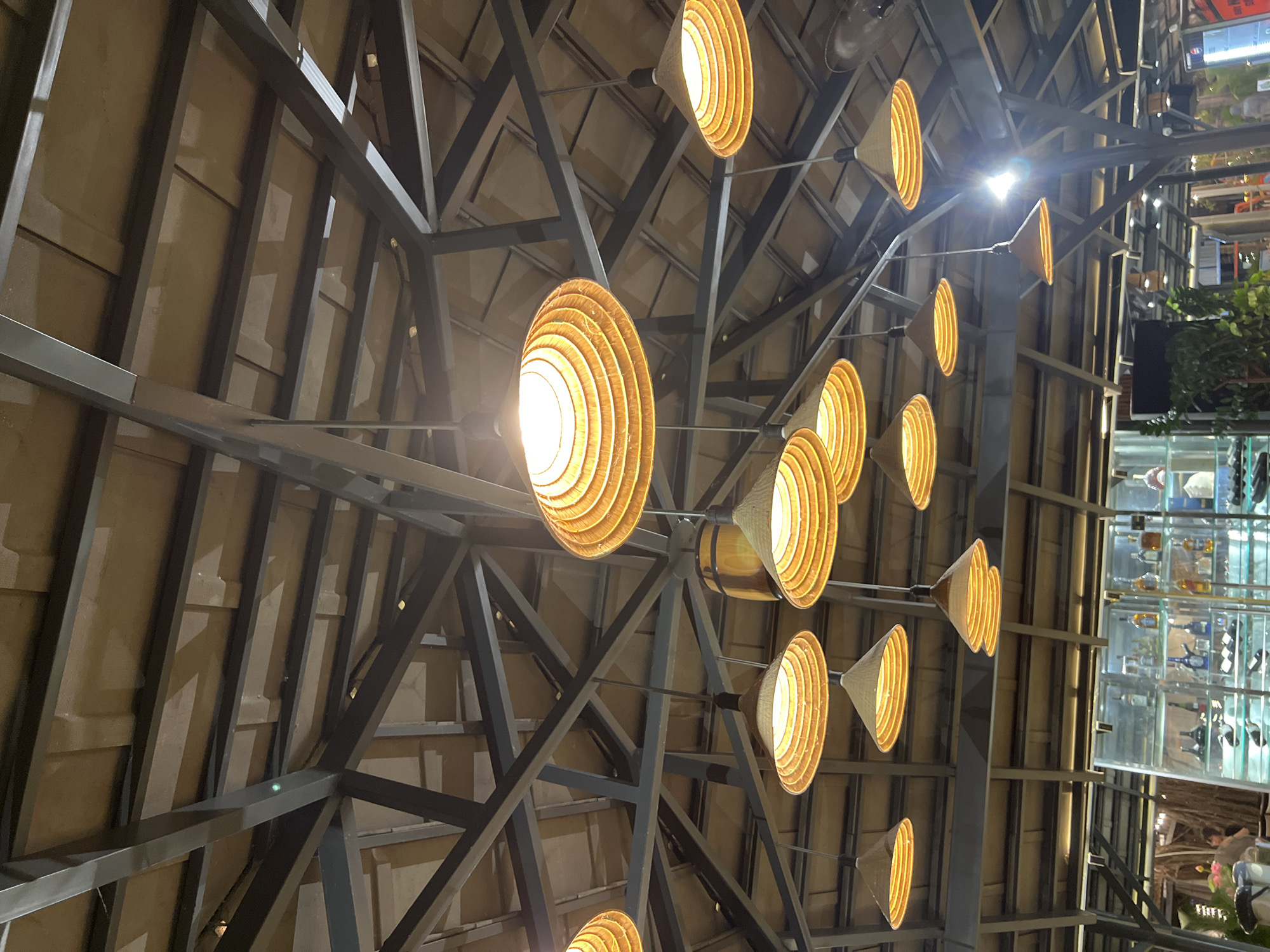 An interior shot captures the ceiling of a restaurant in Bac Nha Trang Ward, Vietnam. The ceiling is constructed with dark metal beams supporting a thatched or woven material, creating a rustic aesthetic. Numerous conical bamboo lamps are suspended from the ceiling, emitting a warm, yellow light. One particularly large, flat, round lamp is prominently displayed in the upper center, while others are arranged in a scattered pattern throughout the frame. A wooden barrel hangs from the ceiling to the left of the large lamp. Below the ceiling, in the background, a glass-fronted display cabinet filled with bottles of alcohol is visible. A few blurred figures can be seen in the lower left and right corners of the image, suggesting the presence of patrons. The overall impression is a dimly lit, atmospheric dining space.