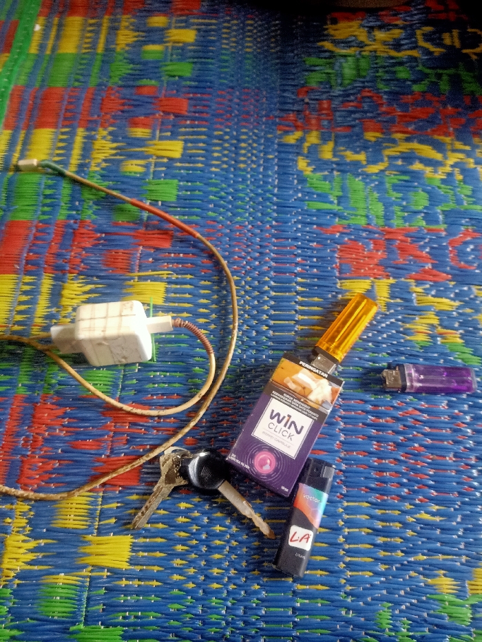 The image displays an arrangement of personal items resting on a brightly colored, woven plastic mat. The mat features a repeating pattern of interwoven blue, red, yellow, and green strands, forming a grid-like texture.

In the upper left, a white power adapter or charger block is visible, connected to a brown and off-white cable that snakes across the mat. The cable appears somewhat worn.

Towards the center-right, a purple pack of "WIN CLICK" cigarettes is prominently placed. The pack has a capsuled filter, indicated by "CLICK" and a circular graphic. Text on the pack includes "PERINGATAN" (Indonesian for "Warning") and other smaller, unreadable text. A yellow-topped lighter is attached or resting on top of the cigarette pack.

Below the cigarette pack and slightly to the left are two keys on a key ring. To the right of the cigarette pack, a purple disposable lighter is lying horizontally. Further down and to the right, a black rectangular device, possibly another lighter or a small vape, is visible. This device has colorful branding that includes the word "Vector" and the letters "LA" and "LYFE" (or similar).

No people are present, and no specific activity is taking place; the items are stationary. The lighting suggests an indoor setting or diffused daylight. The scene appears to be a casual display of everyday objects in a personal space within Pubian, Indonesia.