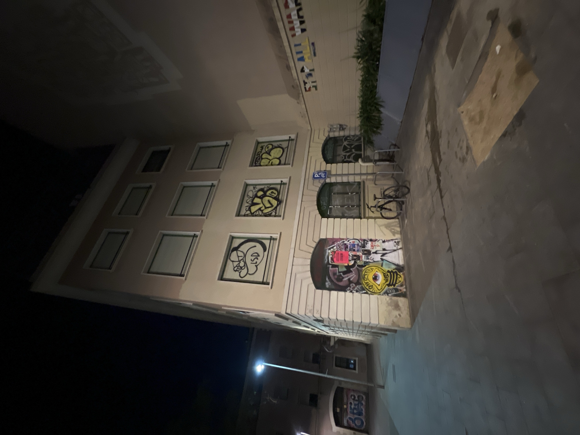 The image shows a building in Barcelona, Spain, at night, with several windows covered by closed shutters. Some of the windows are adorned with graffiti. The building has an arched entranceway on the ground floor, which is also covered in graffiti and posters. In front of the building, there is a bicycle parked, and a blue parking sign with a bicycle symbol. A street lamp is visible in the background, illuminating a street. The ground in front of the building is paved, with some wet patches and a piece of plywood lying on it. The overall scene appears to be a street in a city, possibly in a residential or commercial area, with evidence of urban art and public space usage.