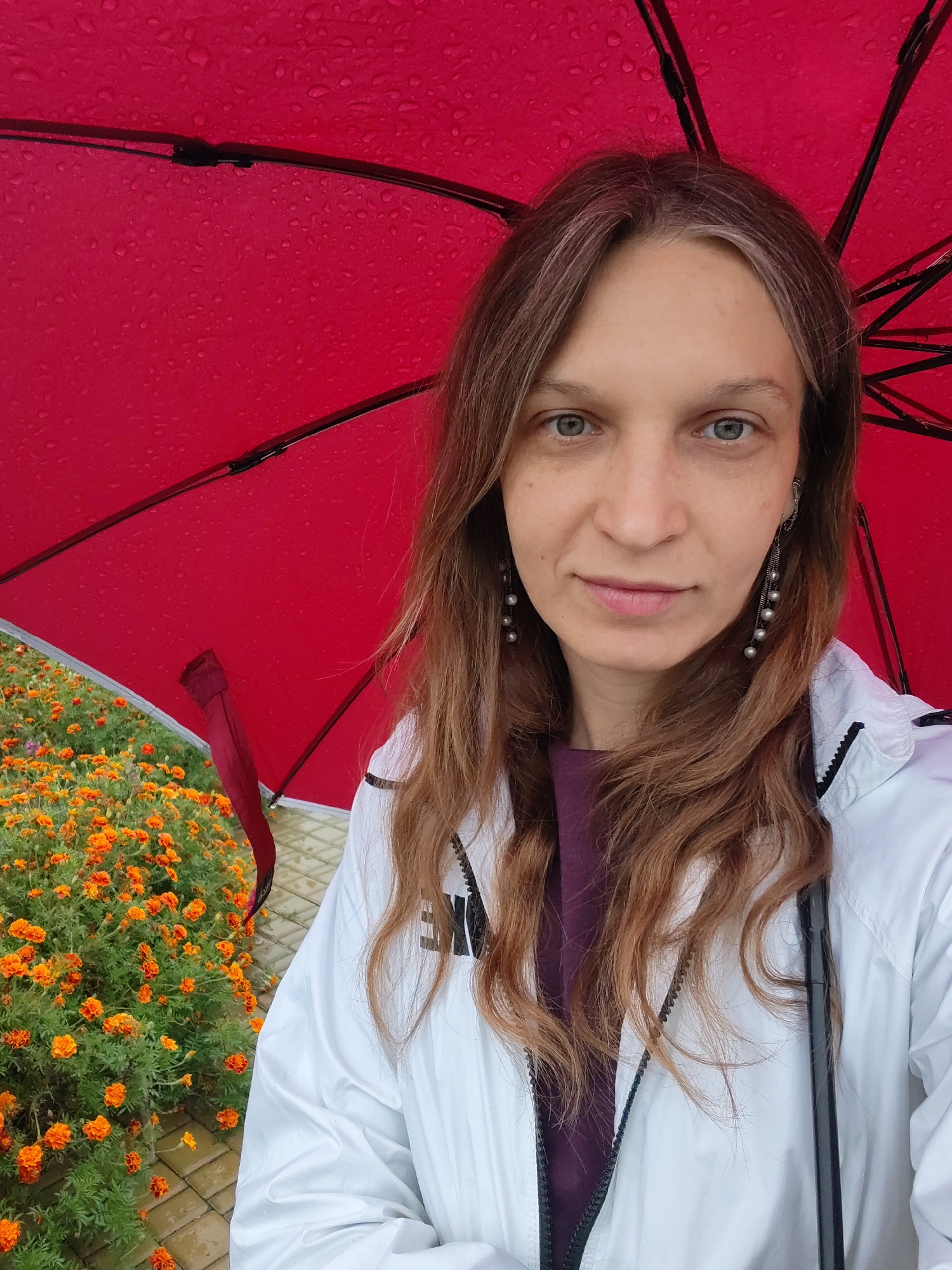 A woman is taking a selfie under a red umbrella. She has shoulder-length brown hair with some gray streaks and is wearing a white jacket over a purple shirt. She has on long pearl earrings. The umbrella is wet with raindrops. To the left of the woman is a patch of bright orange marigolds. The ground is paved with square tiles.