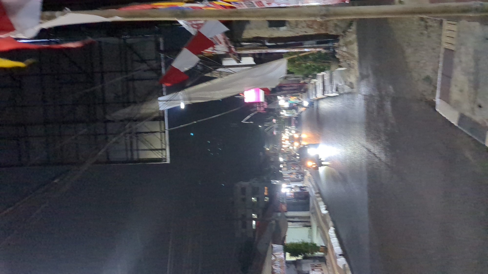 The image depicts a street scene at night in Purwakarta, Indonesia, with vehicles and a pedestrian visible. The street is paved and wet, reflecting the lights of passing vehicles. Buildings line both sides of the street, some with illuminated signs. Overhead, power lines and a large, dark structure are visible against the night sky. Flags, including some with red and white stripes, are hanging from poles on the right side of the image. The overall atmosphere is that of a busy urban environment at night.