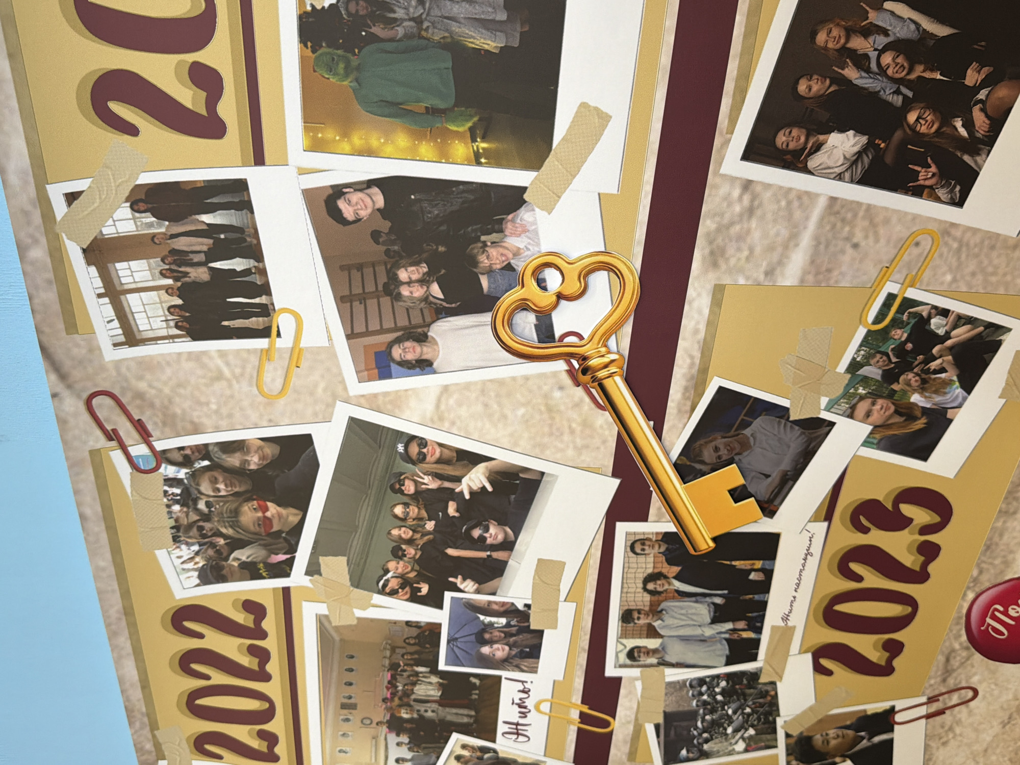 The image shows a collage with the years "2022" and "2023" prominently displayed. It features several photographs of young people, seemingly students, in various group settings. Some photos are attached with tape and paper clips. A large, golden key is centrally positioned over the collage. The collage is set against a pale, marbled-look background, and a portion of blue is visible on the left. There is no information to indicate this is Podolsk, Russia.