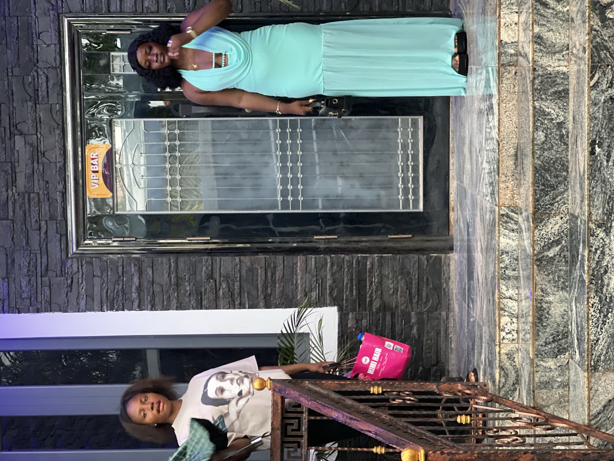 This outdoor photograph captures two women posing at the entrance of a building in Onitsha, Nigeria, likely during the daytime under clear weather conditions. The image, originally rotated, shows the subjects standing on different levels of what appears to be a multi-tiered entrance.
In the foreground, a woman with dark hair wears a light-colored t-shirt featuring a graphic of a person's face. She holds a bright pink bag displaying the text "MIMI HAIR" and other smaller logos, along with what appears to be a mobile phone. She looks towards the camera with a neutral or slightly surprised expression.
Further back, on a higher level, another woman stands in front of a dark, reflective door. She is dressed in a long, flowing light blue (aqua or mint green) dress and holds a small black purse or clutch. Her dark, curly hair frames a smiling face as she too looks towards the viewer. The door behind her has a dark frame, glass panels with an ornate silver design, and a prominent orange sign that reads "VIP BAR".
The building's exterior features a dark, textured stone or brick-like wall. The ground and steps are covered with patterned marble or granite tiles, predominantly in shades of grey and black. A decorative, dark-colored metal railing with intricate designs is visible in the middle ground, separating the two women. Green foliage from plants can be seen in the background, adding a touch of nature to the urban setting. Both women appear relaxed and are actively posing for the photograph.