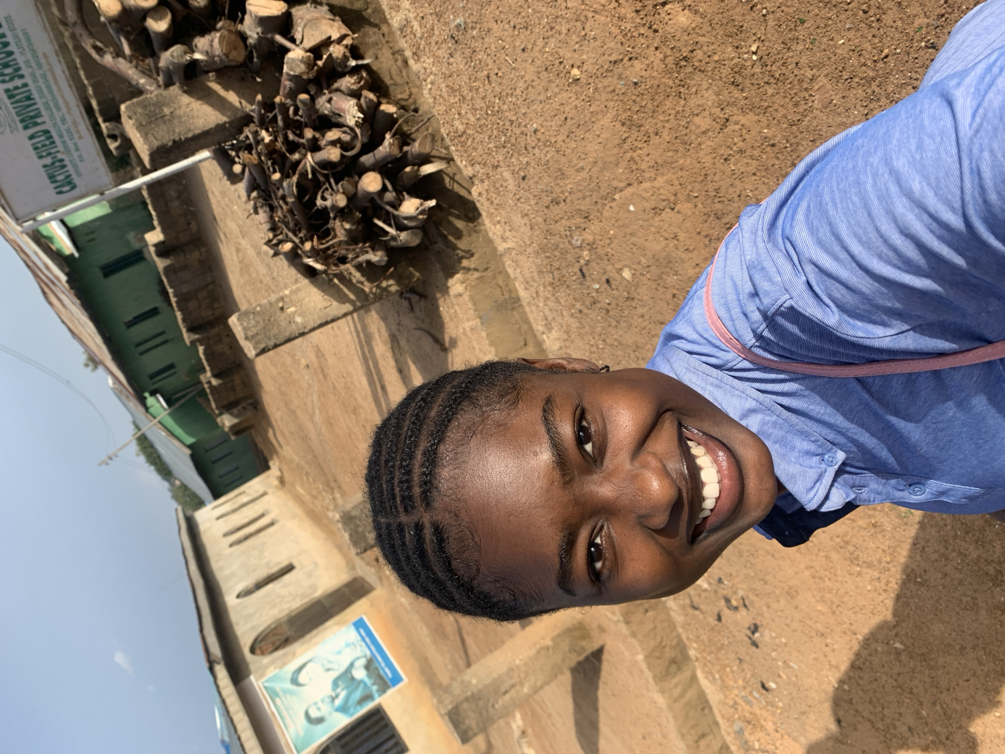 A young woman stands smiling in front of a building with a sign identifying it as "Cactus-Field Private School." She is wearing a blue collared shirt and has a pink strap across her shoulder. Behind her, a pile of cut logs rests against the side of the building. The location is Rafin Par, Nigeria.
