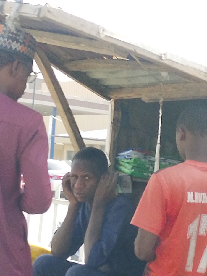 The scene, located in Gandu Albasa, Nigeria, depicts three males near a rudimentary outdoor stall. An adult male is positioned on the left, with his back partially to the camera, wearing a purple long-sleeved shirt and a patterned cap, with glasses visible. In the center, beneath a wooden and corrugated metal shelter, an adult male is seated, facing slightly right and looking towards the camera. He wears a dark blue long-sleeved garment, with his forearms resting on his knees and hands touching the sides of his head. On the right, a young male stands with his back mostly to the camera, wearing an orange short-sleeved t-shirt with "M. MURAD" and the number "17" printed in white on the back. Inside the shelter, stacked green-packaged items are visible on a shelf, along with a circular dark object. In the background, a light-colored multi-story building with horizontal architectural details, a white sedan vehicle, and white metal railings are present. The lighting suggests daytime.