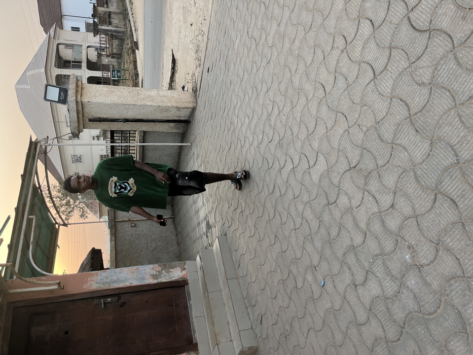 A young man stands on a patterned gray brick walkway. He is wearing a green t-shirt and black shorts. He is holding a black object in his hand. To his left is the corner of a building with a small doorway and steps. Beyond the man is a tall gray stone gatepost and a building with a peaked roof. The sky is light orange, suggesting it is either dawn or dusk.