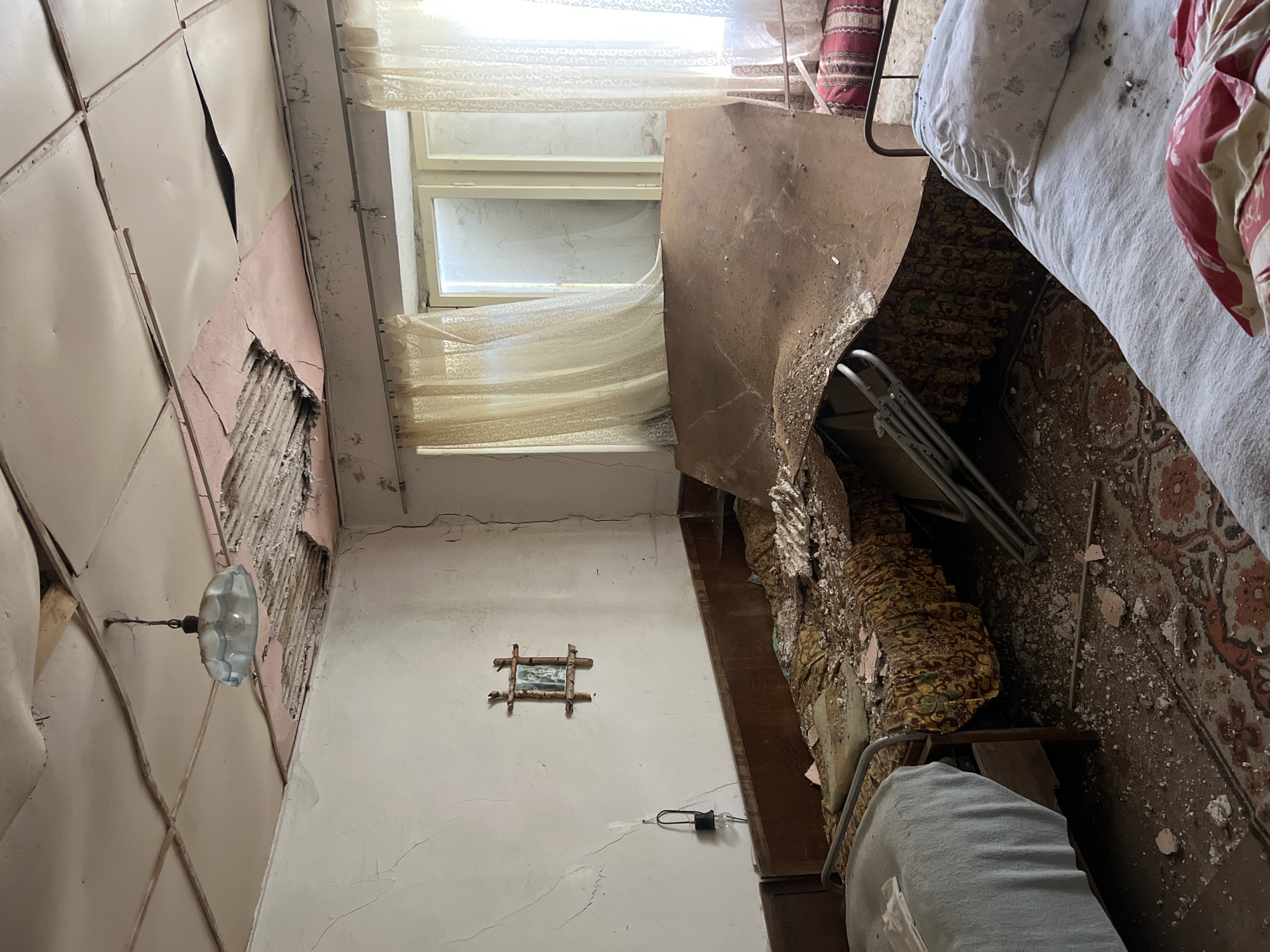 The image shows the interior of a dilapidated building, possibly in Boikovo, Bulgaria. The room is in disrepair with damaged walls, exposed lath, and cracked plaster. A window with lace curtains allows light into the room. Furnishings, including a bed with patterned bedding and a chair, are visible amidst debris on the floor. A framed picture hangs on the wall. A bare lightbulb on a wire is also visible on the wall. Overall, the scene suggests abandonment and deterioration.