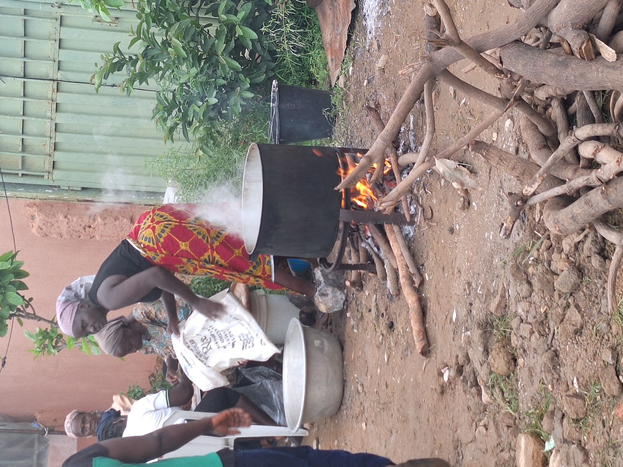In Sabon Tasha, Nigeria, an outdoor scene depicts multiple women preparing food. A woman in a black tank top and a red and yellow patterned skirt pours parboiled rice from a white sack into a large metal basin positioned on the dirt ground. Steam emanates vigorously from a large black pot, centrally placed on a metal stand and heated by an open wood fire. Numerous wooden logs are arranged around and beneath the pot, with additional fuel wood stacked nearby. To the left of the active woman, a silver-colored metal basin rests on the ground. In the background, two other women, one wearing a patterned dress and a head covering, are seated, appearing to handle food items or bags. A fourth individual's arm and leg are partially visible on the far left. The ground is unpaved dirt with scattered small rocks and minimal green growth. The background features a light brown building wall on the left, a light green corrugated metal gate on the right, and dense green foliage. A black plastic bucket is visible near the cooking fire.