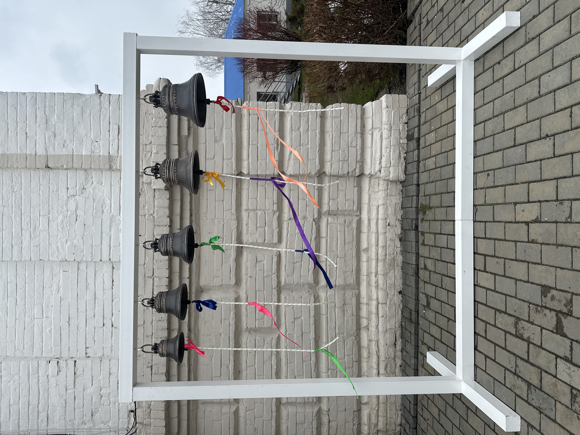 A white rectangular metal frame holding five bells is positioned in an outdoor setting. The bells hang in a vertical line, each connected by a white string to a white brick wall behind the frame. Each bell is tied with a ribbon of varying colors including yellow, orange, purple, blue, red, and green. The background includes a building with a blue roof and bare trees. The ground consists of grey brick pavers. This image likely depicts a public art installation in Votkinsk, Russia.