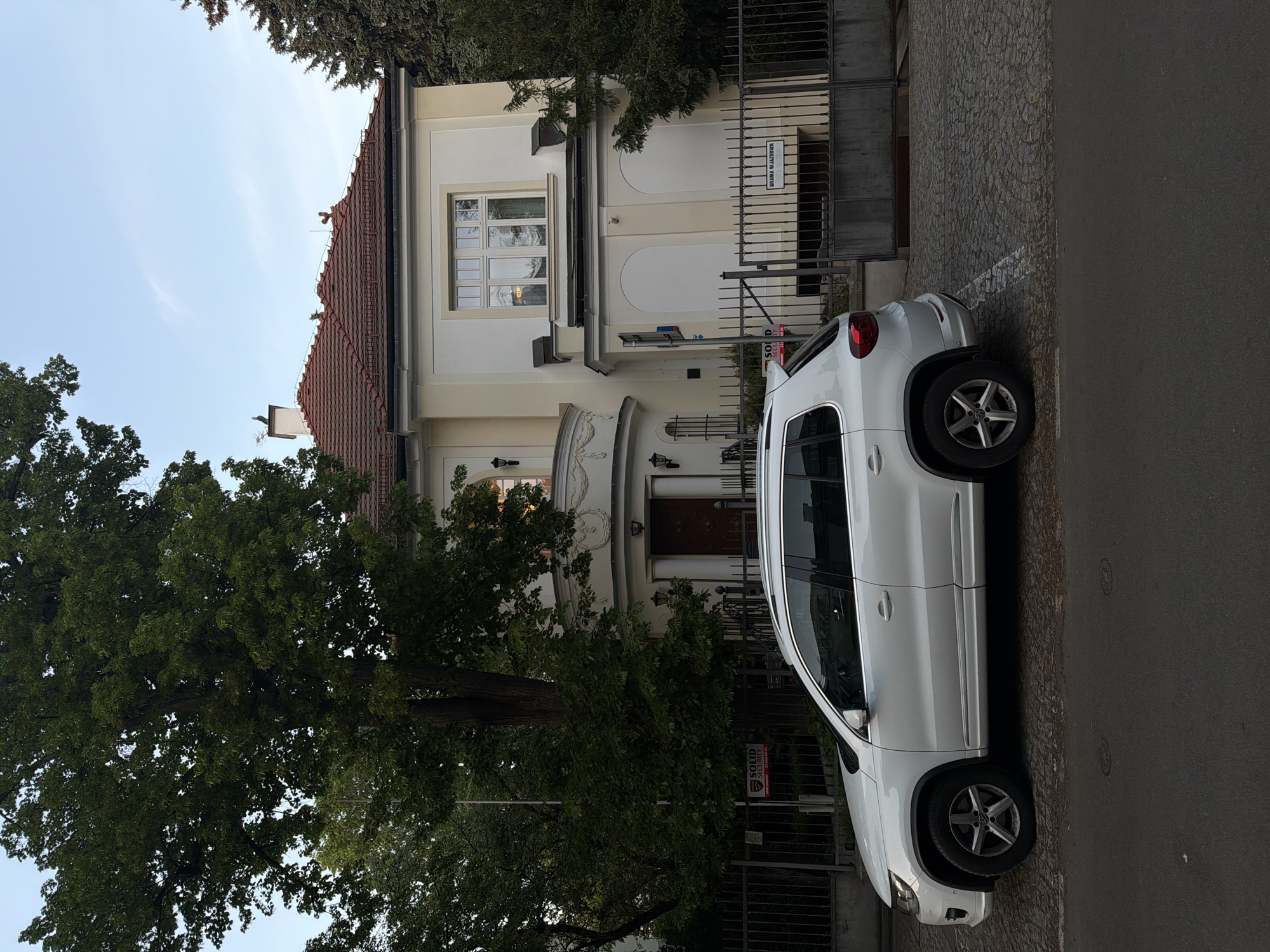 A white SUV is parked on a cobbled street next to a building with a red tile roof. The building is light colored with a decorative facade. A tree partially obscures the left side of the building. A black metal fence is located between the car and the building. A sign in a foreign language is visible near the entrance to the building. The street is paved with asphalt. The sky is clear and light blue. A security system sign is posted on the fence. The location is Unknown, Unknown.