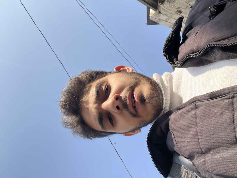 The image presents a low-angle, head-and-shoulders portrait of a young adult male against a clear blue sky. The individual has dark hair, a short beard, and a mustache. He is wearing a white ribbed turtleneck sweater and a dark, possibly black, quilted jacket. A grey backpack strap is visible over his left shoulder. His mouth is slightly open, and he is looking directly into the camera. Two dark utility wires traverse the upper portion of the frame against the sky. A partial view of a light-colored building structure with a darker roofline is visible on the far right edge. The scene is illuminated by bright daylight and is located in Rukn ad-Din Municipality, Syria.