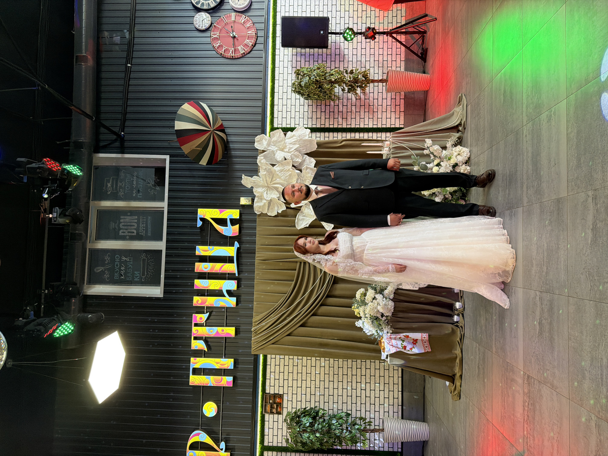 A wedding photo is taken of a bride and groom standing side-by-side in front of a draped background. The bride is wearing a white lace wedding dress and a veil, while the groom is in a dark suit and tie. They are holding hands. The room appears to be a banquet hall with decorative elements such as illuminated letters on the wall, large paper flowers, clocks, and a decorative umbrella. There are also potted plants, a speaker on a stand, and colored stage lights casting green and red hues on the floor. The floor is tiled. The lettering on the wall is in Russian and likely spells out "Свадьба" (Wedding) or a similar celebratory phrase. The presence of multiple clocks on the wall, particularly the large red-faced one, could be a reference to time or a nod to the city's history if it's a local landmark. The overall impression is a festive and decorated wedding reception venue, possibly in Chernushka, Russia, given the Cyrillic lettering.