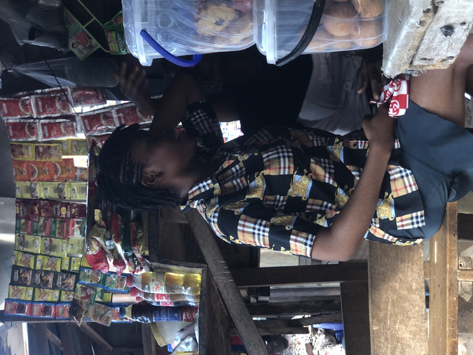 A woman sits, turned to the left, near a stand filled with small goods in red and yellow packaging. She wears a checkered shirt with images of horses and is holding something in her hands. Above the stand are transparent plastic containers filled with baked goods. Someone stands to the right of the woman wearing white shorts, and to her left, someone appears to be holding a microphone. The setup appears to be outdoors.