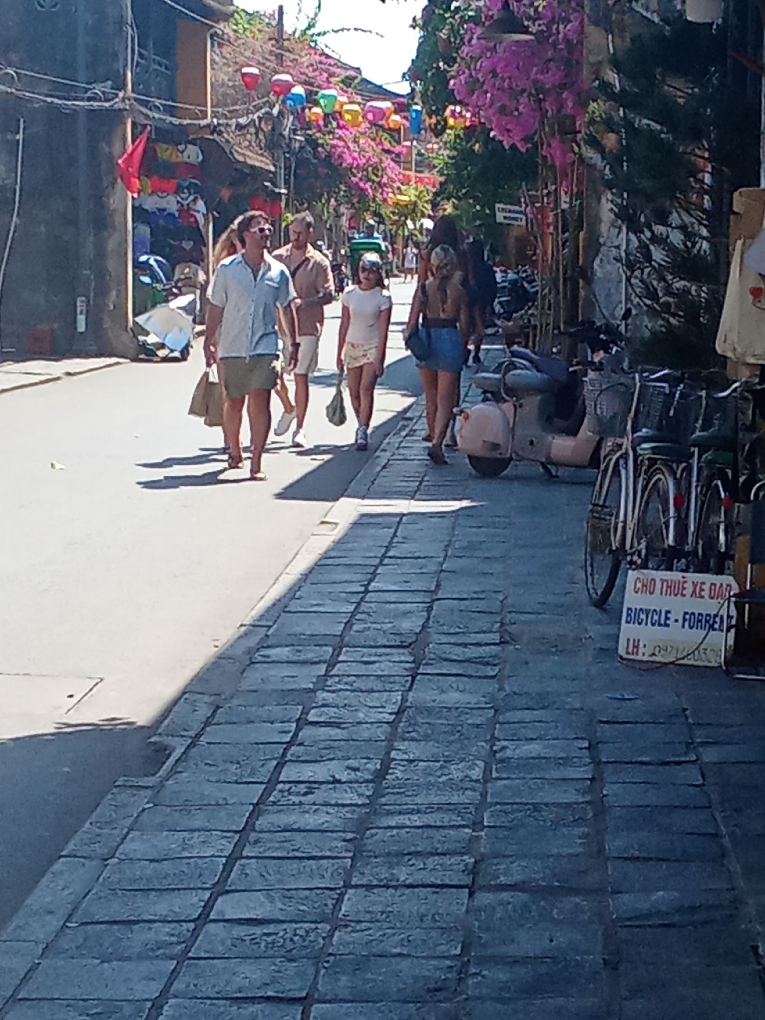 The image depicts a street scene in Hoi An Ward, Vietnam, likely a tourist area, characterized by a narrow street lined with buildings and adorned with vibrant flowers. A group of people are walking along the cobblestone sidewalk. In the foreground, a man wearing sunglasses, a light blue short-sleeved shirt, and khaki shorts is walking with a brown shopping bag in his left hand. To his right, another man in a light brown short-sleeved shirt and white shorts is walking. Behind them, a woman in a white t-shirt and a patterned skirt is walking, carrying a similar brown shopping bag. Further back, several other individuals are visible, some appearing to be tourists.
On the left side of the street, shops are visible with merchandise displayed. One shop has a red flag hanging. The street is decorated with colorful lanterns strung overhead. In the background, dense pink bougainvillea flowers cascade down from above, adding to the picturesque atmosphere.
To the right of the sidewalk, several parked motorbikes and bicycles are present. One of the bicycles has a sign attached to its basket that reads "CHO THUÊ XE ĐẠP BICYCLE - FOR RENT" followed by "LH: 0971403528". The architecture suggests a historic district, with textured buildings and a paved walkway. The lighting indicates it is daytime with bright sunlight casting shadows on the ground.