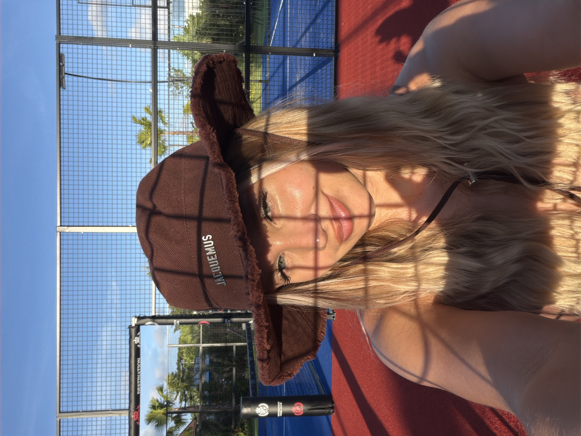 The image depicts a young woman wearing a brown bucket hat with the word "JACQUEMUS" embroidered on it, and a fuzzy brim. She is in an outdoor setting that appears to be a sports court, possibly for padel tennis, with a blue surface and red outer areas. The court is enclosed by a wire fence, and palm trees are visible in the background, suggesting a tropical or subtropical location. The sky is clear and blue, with the sun casting strong shadows across the scene, including on the woman's face. The woman has blonde hair and is looking towards the camera with a smile. There is a black cylindrical object with logos on it near the fence on the left side of the frame. The overall lighting suggests it is late afternoon or early morning. The location is identified as Thep Krasatti, Thailand.