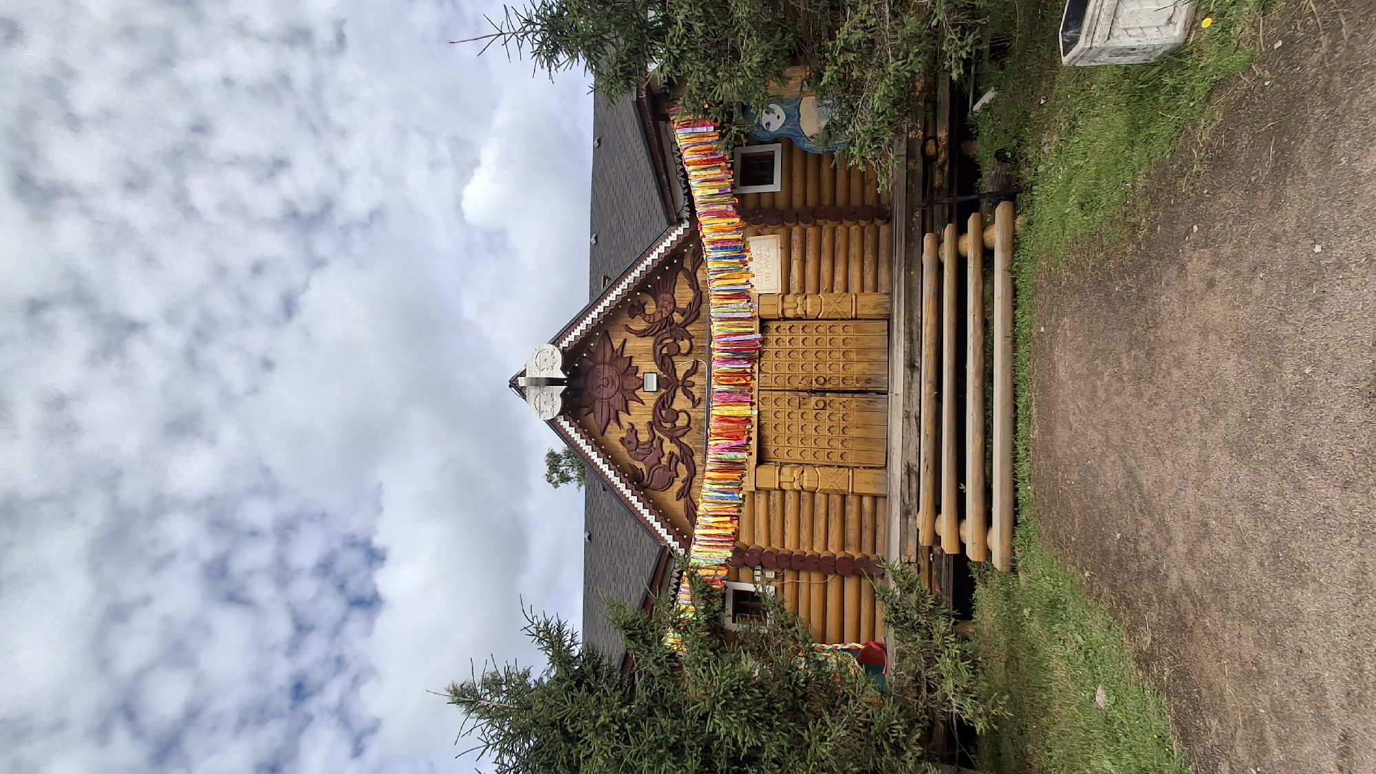 A traditional wooden log building, likely a museum or cultural center, is situated in Peterhof, Russia. The structure features double wooden doors, decorative carvings of mythological creatures and a sun on its facade, and a festive string of colorful ribbons draped above the entrance. It is flanked by evergreen trees and set against a cloudy sky. A wooden staircase leads up to the entrance. The building is set back from a dirt path with grass on either side. A decorative figure, resembling a blue-and-white bear, is visible on the right side of the building.