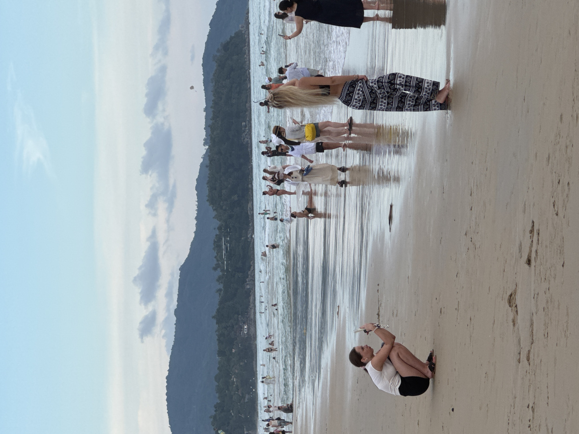 The image depicts a crowded beach scene in Karon, Thailand. In the foreground, two women are visible. The woman on the left is crouching on the sand, holding a smartphone and appearing to take a photo. She is wearing a white tank top and black shorts. To her right, a woman with long blonde hair stands facing the water, wearing a black bikini top and patterned pants. She is also holding a smartphone, possibly taking a photo or video.
The middle ground of the image is occupied by numerous people wading in the ocean and walking along the shoreline. The water is moderately wavy, and several individuals are swimming or standing in the surf. The beach itself is sandy and wet near the water's edge, reflecting the sky.
In the background, a densely forested hillside rises, dotted with buildings, indicating a developed coastal area. The sky is overcast with pale blue and white clouds. Far in the distance, a small parasail is visible in the sky above the water and the hills. The overall atmosphere suggests a popular tourist destination during the day.