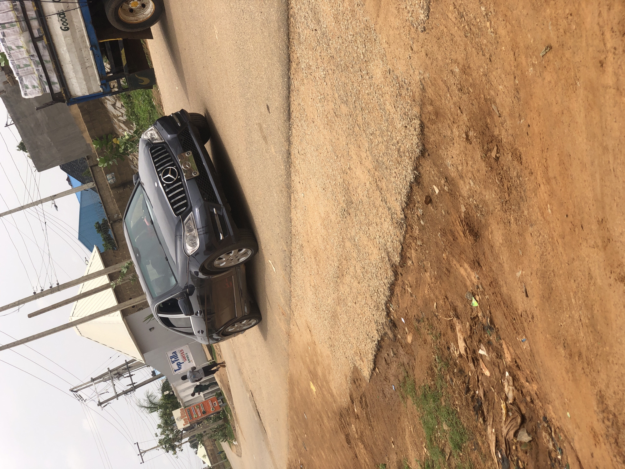 The image depicts a street scene in Hanwa, Nigeria. A black Mercedes-Benz SUV is parked on the side of the road, partially obstructing the lane. Adjacent to the car, a truck with a white bed is also parked. On the opposite side of the street, a building is visible with a sign that reads "Top Villa Suites Annex." A person is walking on the sidewalk in front of the building. Overhead, power lines crisscross the sky. The road is paved but shows signs of wear and tear. To the right of the pavement, there is unpaved terrain consisting of gravel and bare earth.