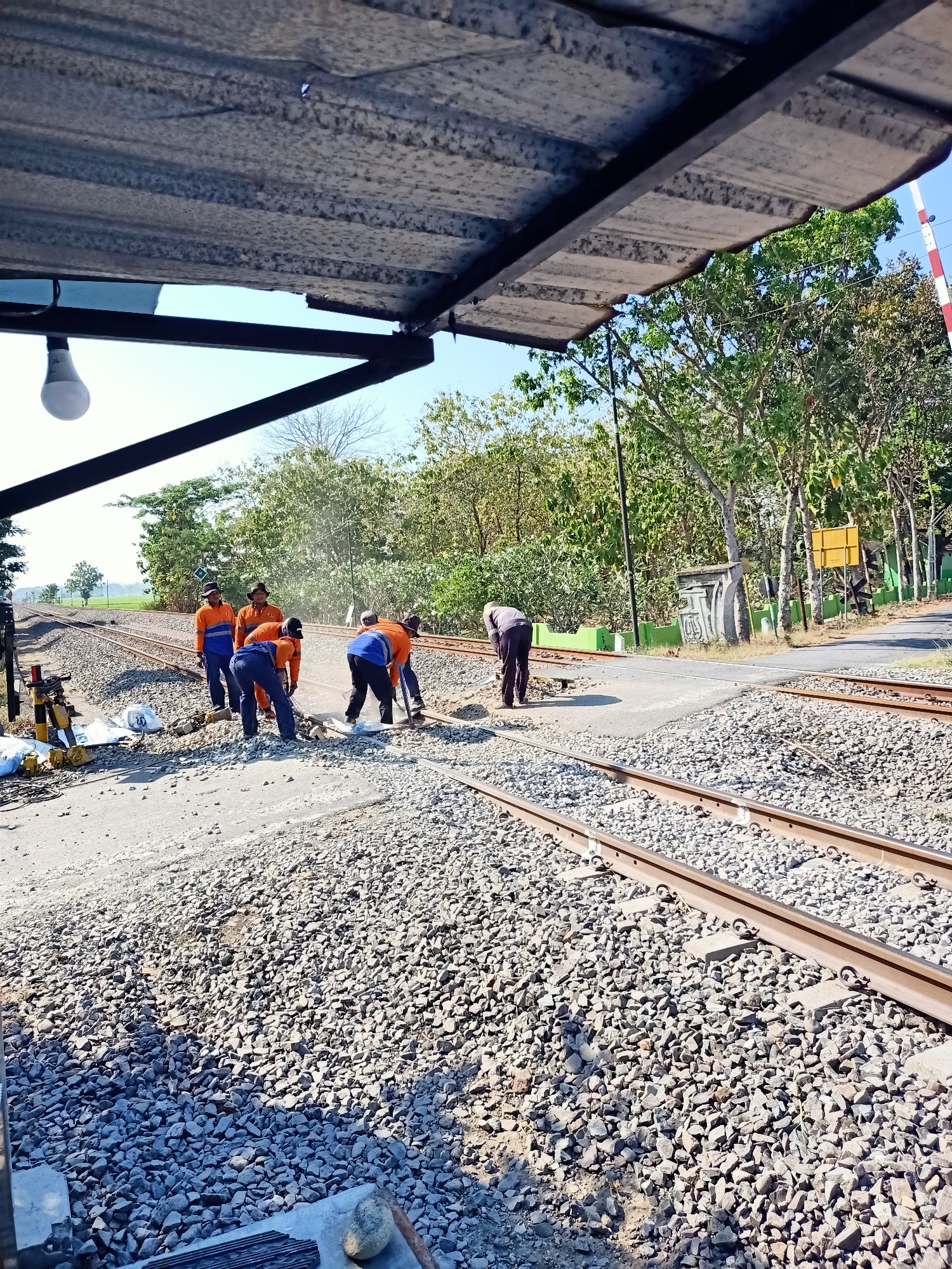 In the city of Banjarjo, Indonesia, a group of workers are engaged in manual labor on railway tracks, possibly undergoing maintenance or construction. The scene is outdoors, under a clear blue sky, with lush green trees and vegetation forming a backdrop. The foreground is dominated by railway tracks and ballast stones, indicative of a railway environment. Several workers, dressed in orange work uniforms and blue pants, are actively involved in shoveling and moving materials around the tracks. Their actions suggest a coordinated effort to manage the railway infrastructure. The presence of a lamp hanging from a covered structure overhead indicates a functional area, possibly a shelter or a maintenance shed. The overall impression is one of diligent work in an industrial or infrastructure development setting within a rural or semi-rural landscape.