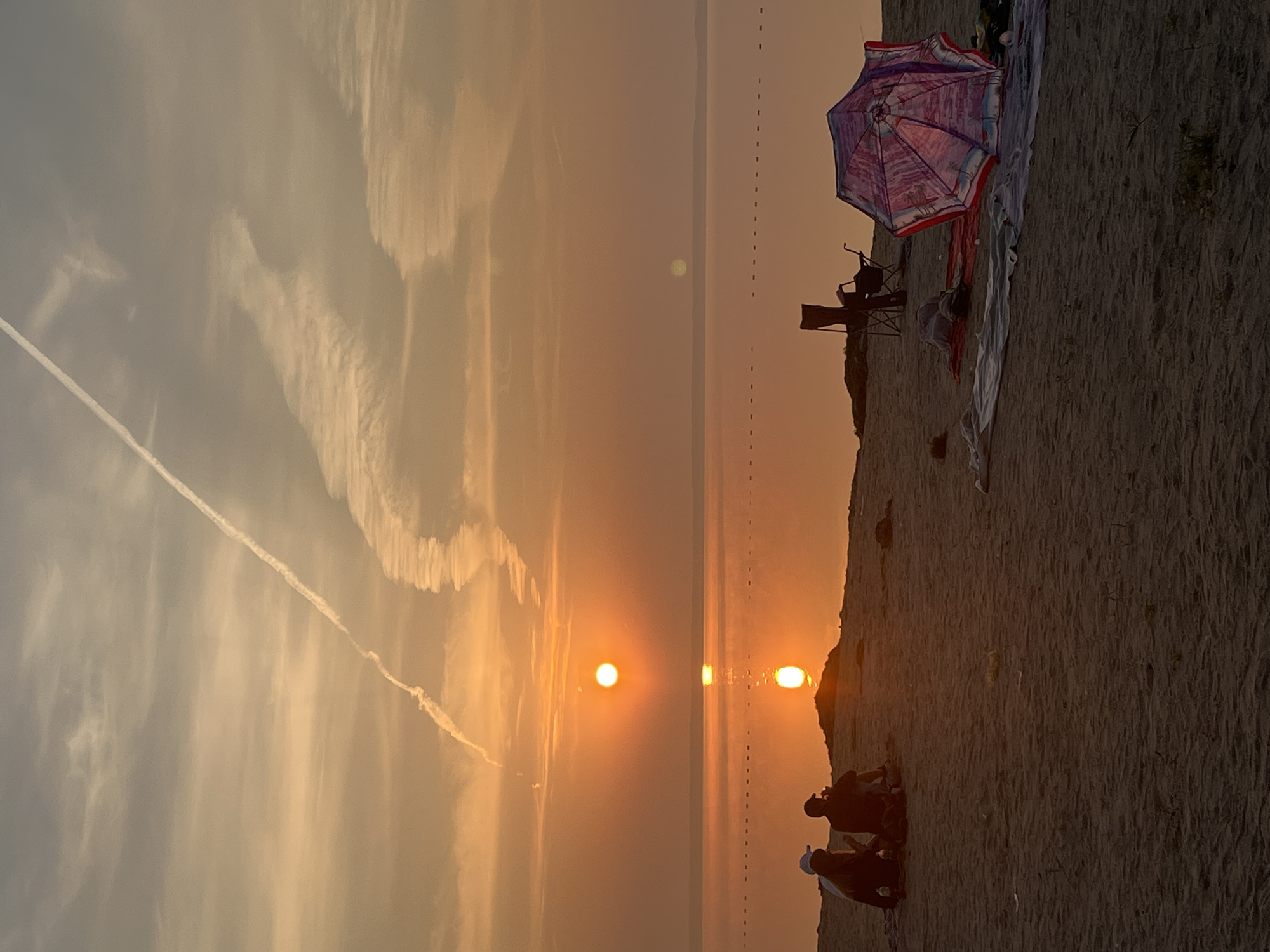 The image depicts a sunset scene on a sandy beach, likely in Surakovo, Russia, given the context. The sun, a bright orange orb, is low on the horizon, casting a warm, golden glow across the sky and water. A distinct contrail streaks across the sky, indicating aircraft activity. In the foreground, two individuals are seated on the sand, facing the water, appearing to be engaged in conversation or enjoying the view. Nearby, beach paraphernalia, including a colorful umbrella, blankets, and chairs, are set up, suggesting a leisurely outing. The water stretches out to the horizon, reflecting the sunset's colors and appearing calm. A series of small buoys or markers can be seen as a dotted line across the water. The sandy terrain of the beach dominates the foreground, with subtle textures and shadows.